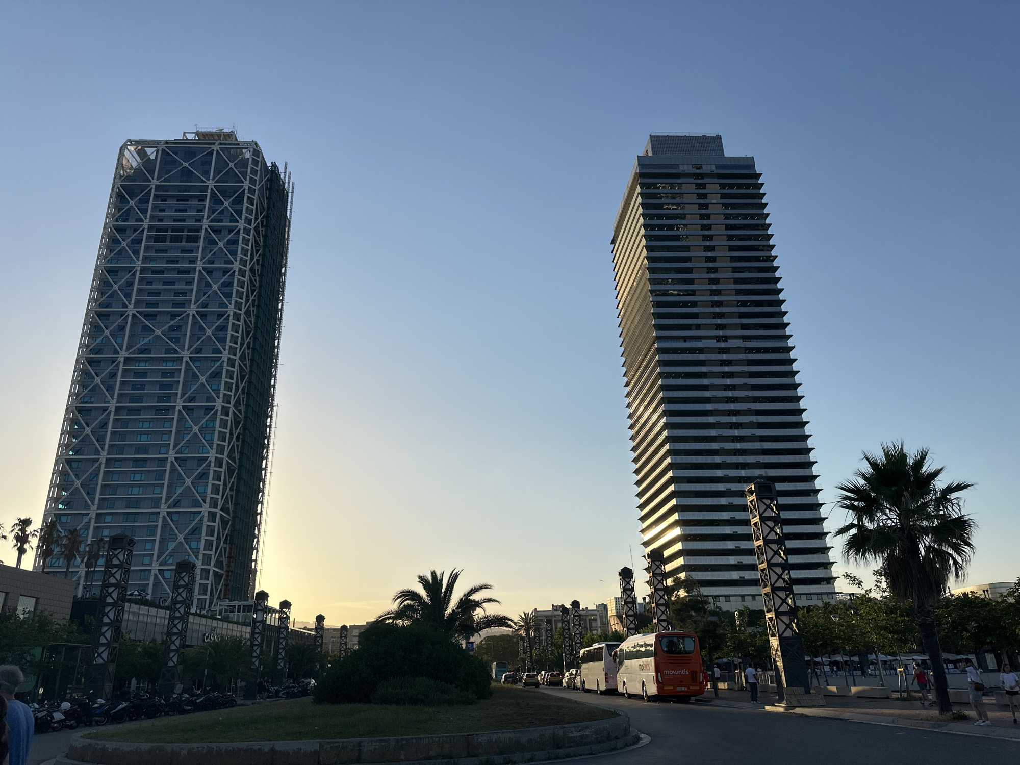 The image captures a view of the Barcelona skyline at dusk, featuring two prominent skyscrapers. The skyscraper on the left has a distinctive white latticework exoskeleton surrounding a glass facade. The skyscraper on the right is a sleeker, dark glass building with visible horizontal divisions and balconies. In the foreground, a road curves around a circular grassy median with several palm trees and bushes. A white bus with "Moventis" branding is visible on the right side of the road, alongside other vehicles in traffic. Parked motorcycles line the sidewalk in front of a building with a sign that appears to say "CASINO". The sky is a clear, pale blue transitioning to a warm yellow hue near the horizon, indicating the setting sun. Several palm trees are scattered throughout the scene, contributing to the Mediterranean atmosphere of Barcelona.