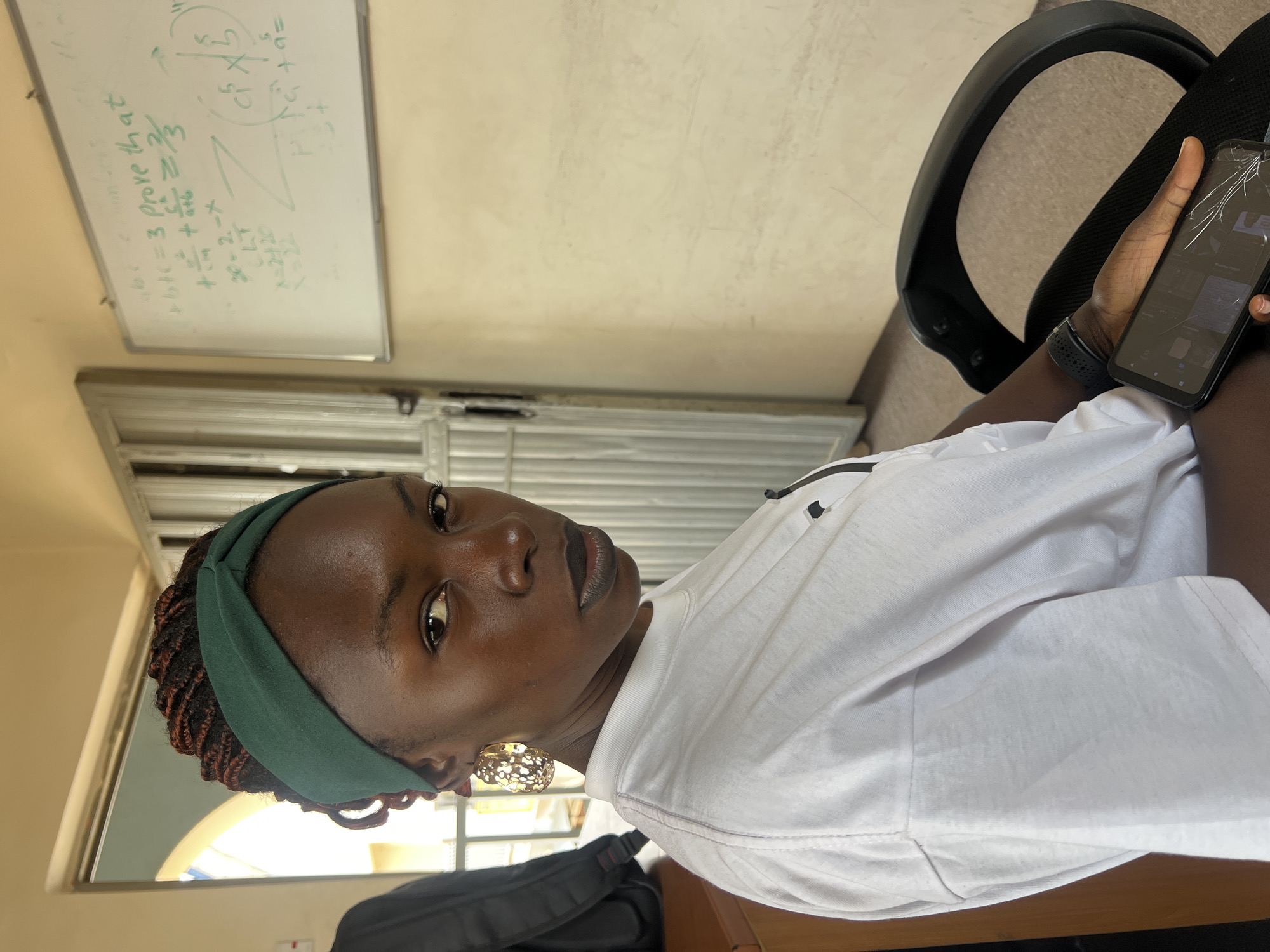 A young Black woman with braided hair and a green headband is seated in a chair, looking directly at the camera. She is wearing a white t-shirt and large gold hoop earrings. A smartphone with a cracked screen rests in her hand. In the background, a whiteboard with mathematical equations is visible on the wall. The scene appears to be an indoor setting, possibly a classroom or office, with a doorway and part of a backpack visible. The lighting is natural and somewhat diffused, suggesting daylight.