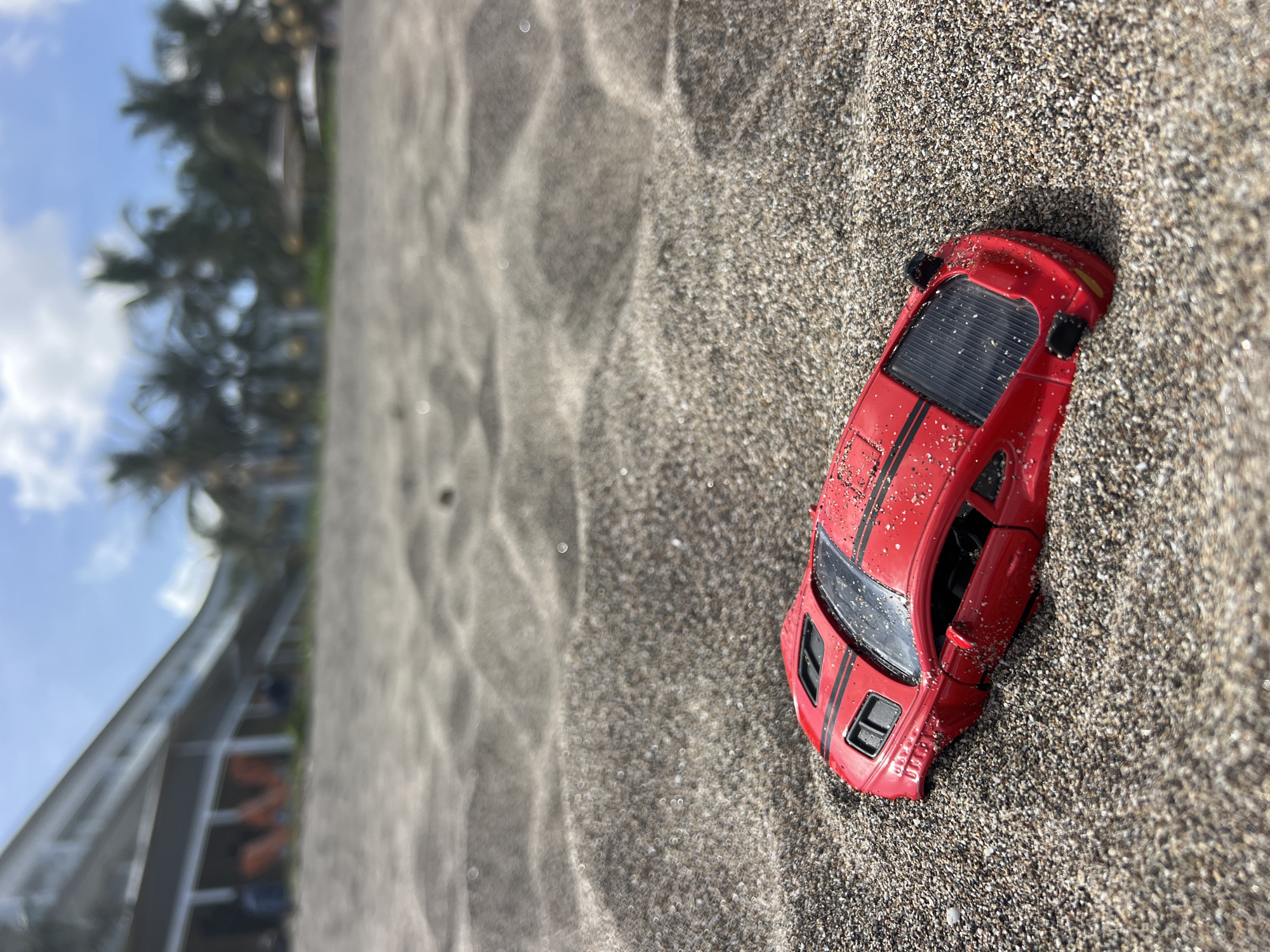 A red toy car with black racing stripes is half-buried in dark sand on a beach. The car is positioned in the foreground and is slightly angled. In the background, out of focus, is a building with a white roof and pillars, and palm trees against a blue sky with white clouds. The setting is a beach in North Kuta, Indonesia. There are no people visible in the immediate vicinity of the toy car.