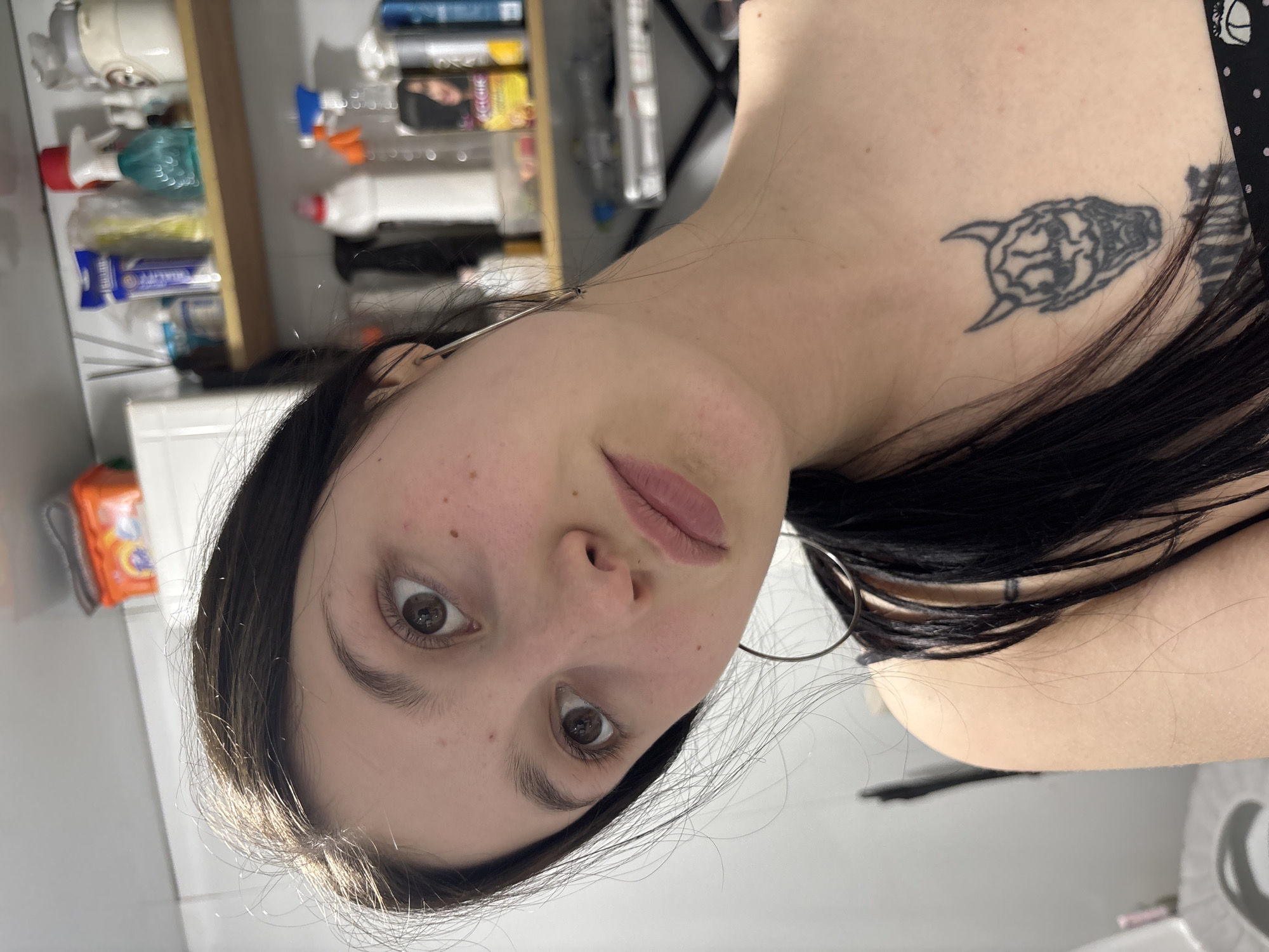 A close-up, slightly tilted photograph captures a young woman with long, dark hair and brown eyes. She wears large silver hoop earrings and has a tattoo on her upper chest, depicting a horned creature. Her skin shows some freckles and minor blemishes.
The background reveals a bathroom setting. Visible elements include a shelf filled with various household cleaning products and toiletries, a white tiled wall, and a glimpse of a toilet.