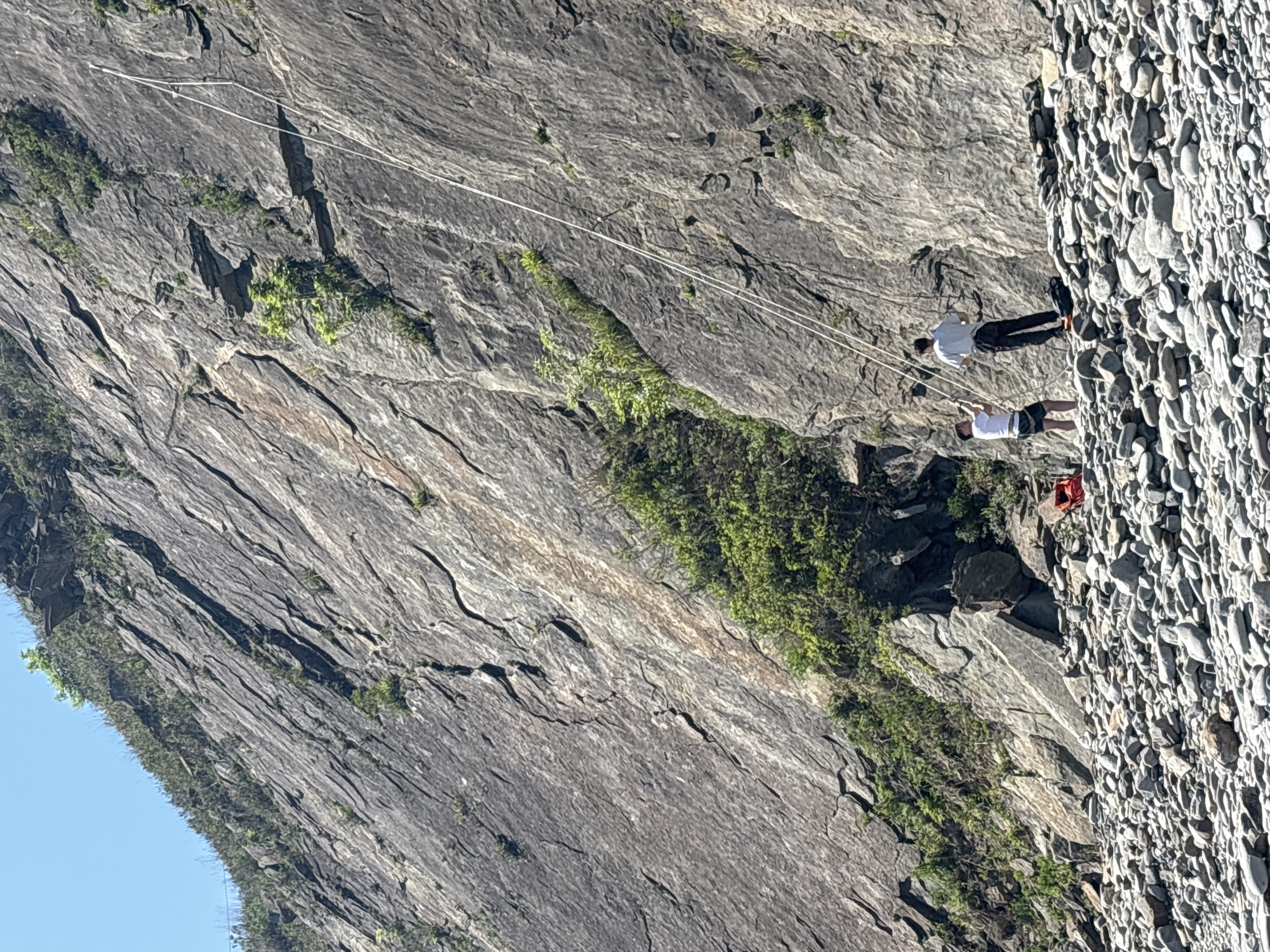 Two people are traversing a rock face. One person is wearing a white shirt and dark pants and the other is wearing a white shirt and dark shorts. They are secured by ropes. The rock face is adjacent to a large, steep embankment of pebbles. The scene appears to be in a rocky, mountainous environment. It is known that Vladivostok, Russia, is located in a mountainous area on the coast of the Sea of Japan.