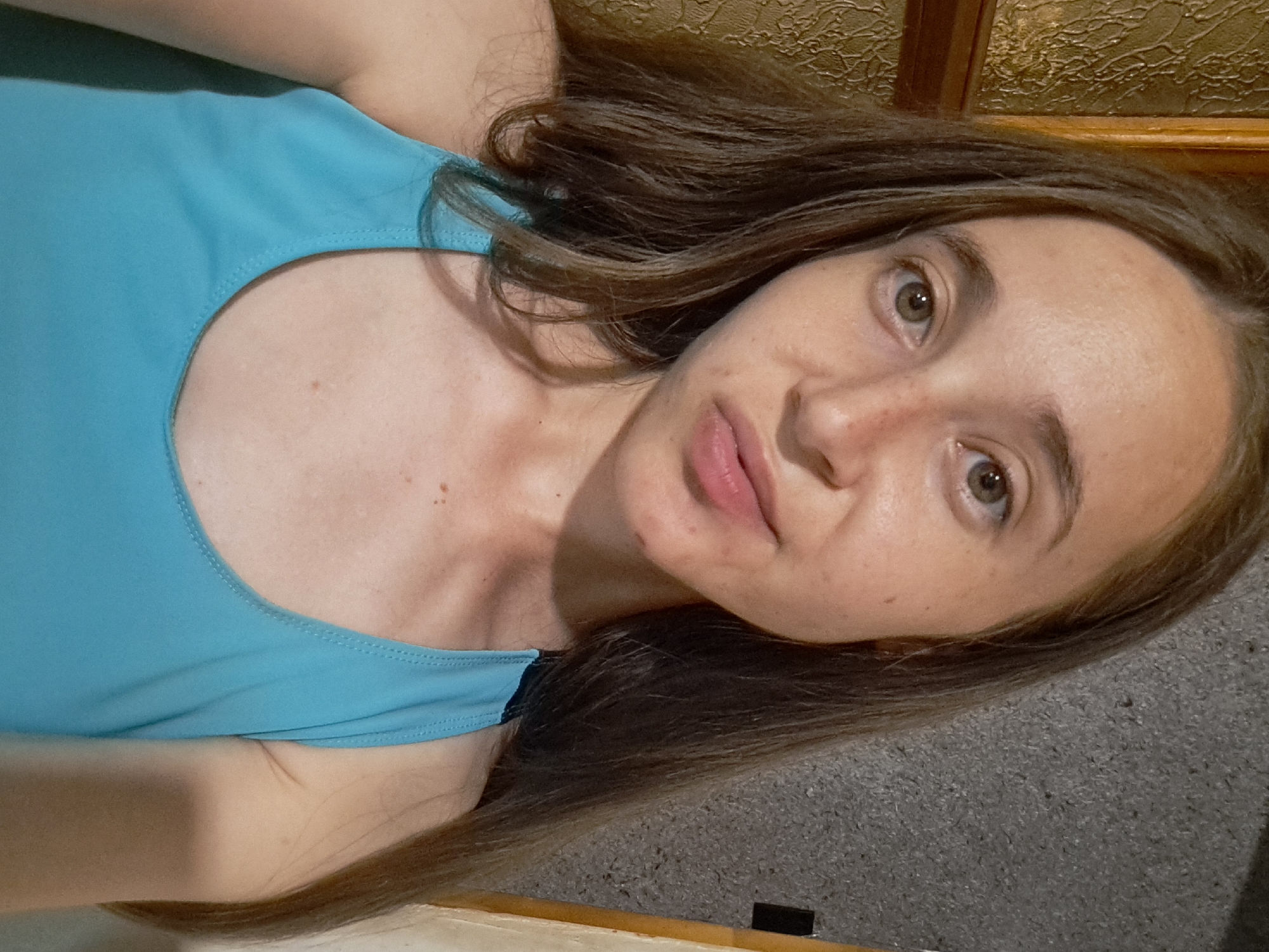 A close-up selfie of a young woman with long, brown hair and freckles on her face. She is wearing a blue tank top. She is indoors, with a textured grey wall behind her and a wooden-framed object to her left. The lighting suggests it is daytime. The scene appears to be within a residential setting.