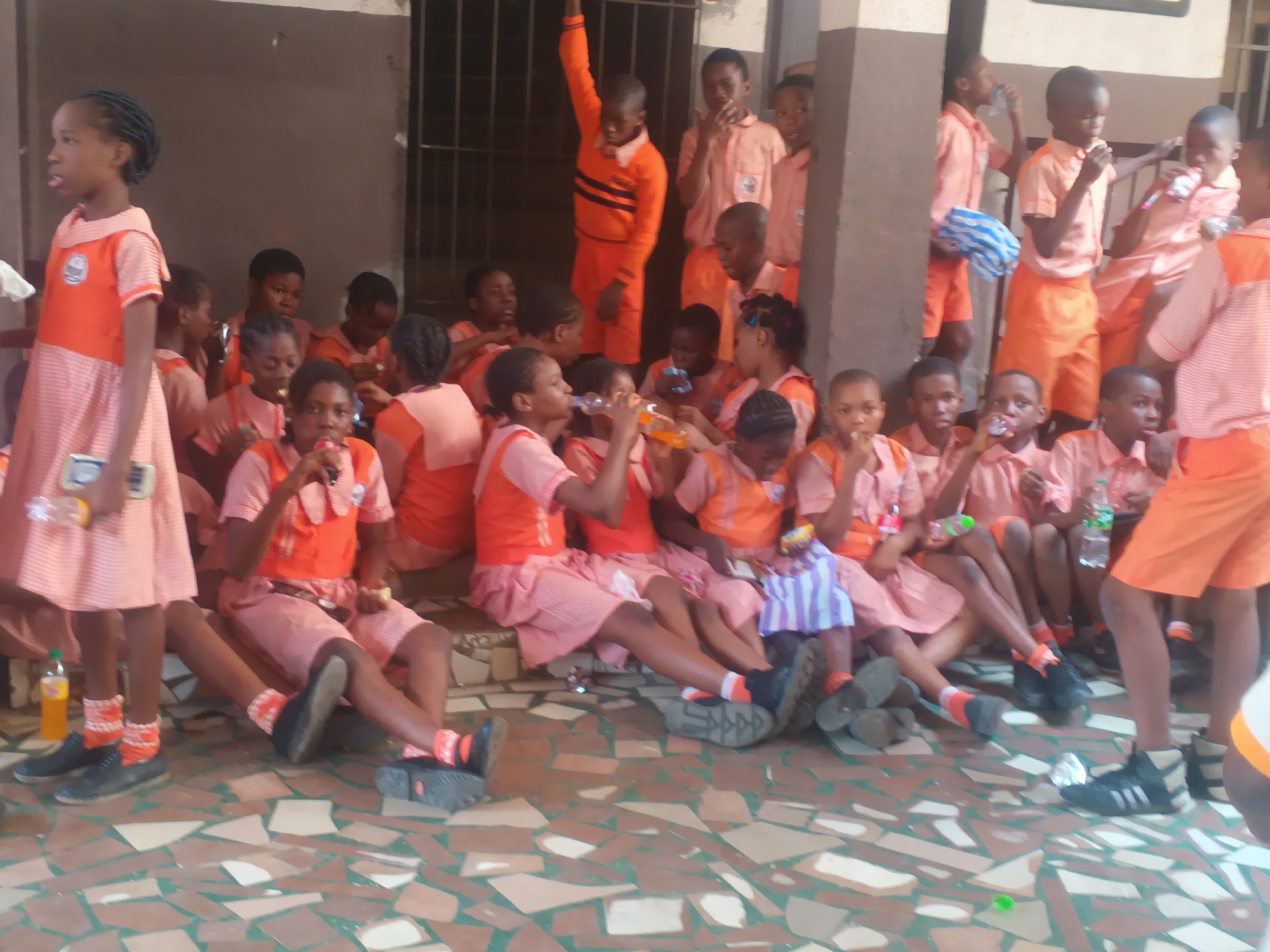 The image depicts a group of Nigerian schoolchildren, predominantly wearing orange and white uniforms, gathered outdoors. The setting appears to be a school or community area in Onitsha, Nigeria, characterized by a building with brown walls and a barred window in the background, and a tiled, patterned ground in the foreground. The children, ranging in age, are seated and standing, many holding or drinking from bottles. Some are interacting with each other, while others appear to be listening or observing. The overall atmosphere suggests a break or a gathering during a school day.