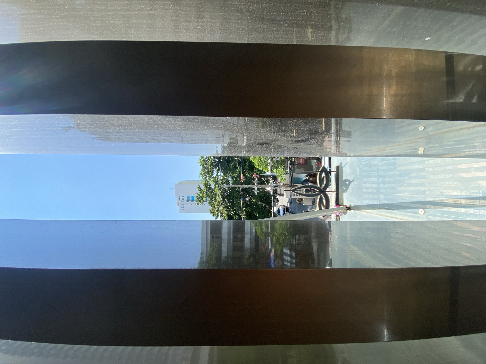 The image displays a view through a modern art installation or architectural feature composed of vertical, dark, reflective metallic panels and transparent panels. The foreground is dominated by these panels, which create a layered, framed perspective of an outdoor urban scene. Through the gaps between the panels, a clear blue sky is visible above a street. In the mid-ground, several buildings of varying heights are present, including a white high-rise with blue-tinted windows. Lush green trees line the street, partially obscuring the buildings. A street scene unfolds below, with a car, traffic lights, and what appears to be a street intersection. A decorative metallic element with a symmetrical, flowing design is situated in the lower-middle portion of the frame, resting on a transparent surface. The overall impression is a contemporary urban environment in Nha Trang Ward, Vietnam, captured from a unique vantage point within a modern structure.