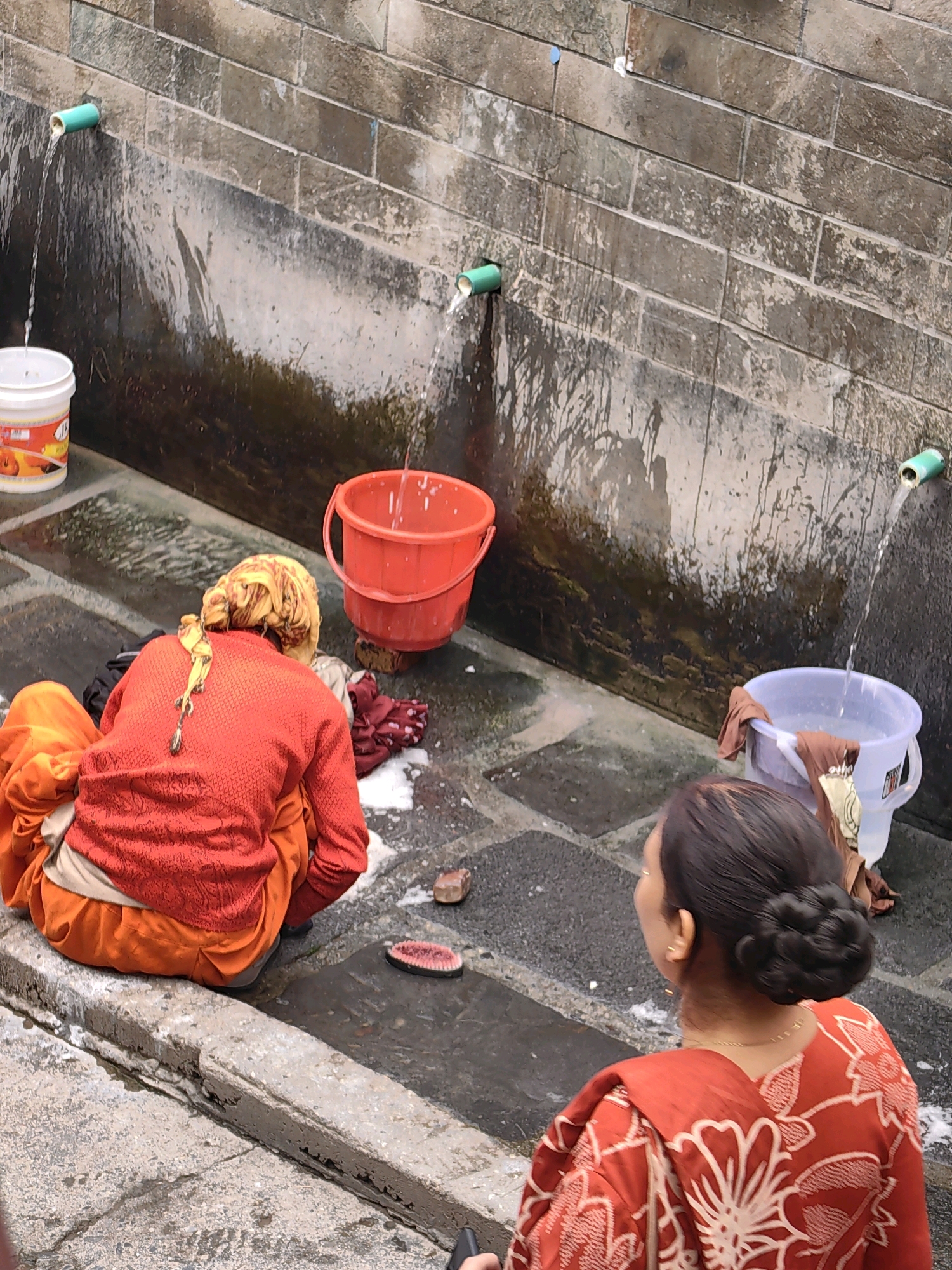 In the city of Vashisht, India, two women are engaged in laundry activities next to a stone wall. The wall has several green pipes protruding from it, from which water flows into buckets and onto the ground. In the foreground, a woman wearing a red garment with a white floral pattern is facing away from the camera, with her hair styled in a bun. To her left, another woman, also facing away, is wearing a red top and orange lower garment, with her hair covered by a yellow scarf. She is seated next to a pile of clothes. Several buckets, one red and two white/clear, are present, one of which contains water and a piece of clothing. The ground is paved with stone tiles, and water is running in channels. A slice of watermelon and a small stone are also visible on the ground.