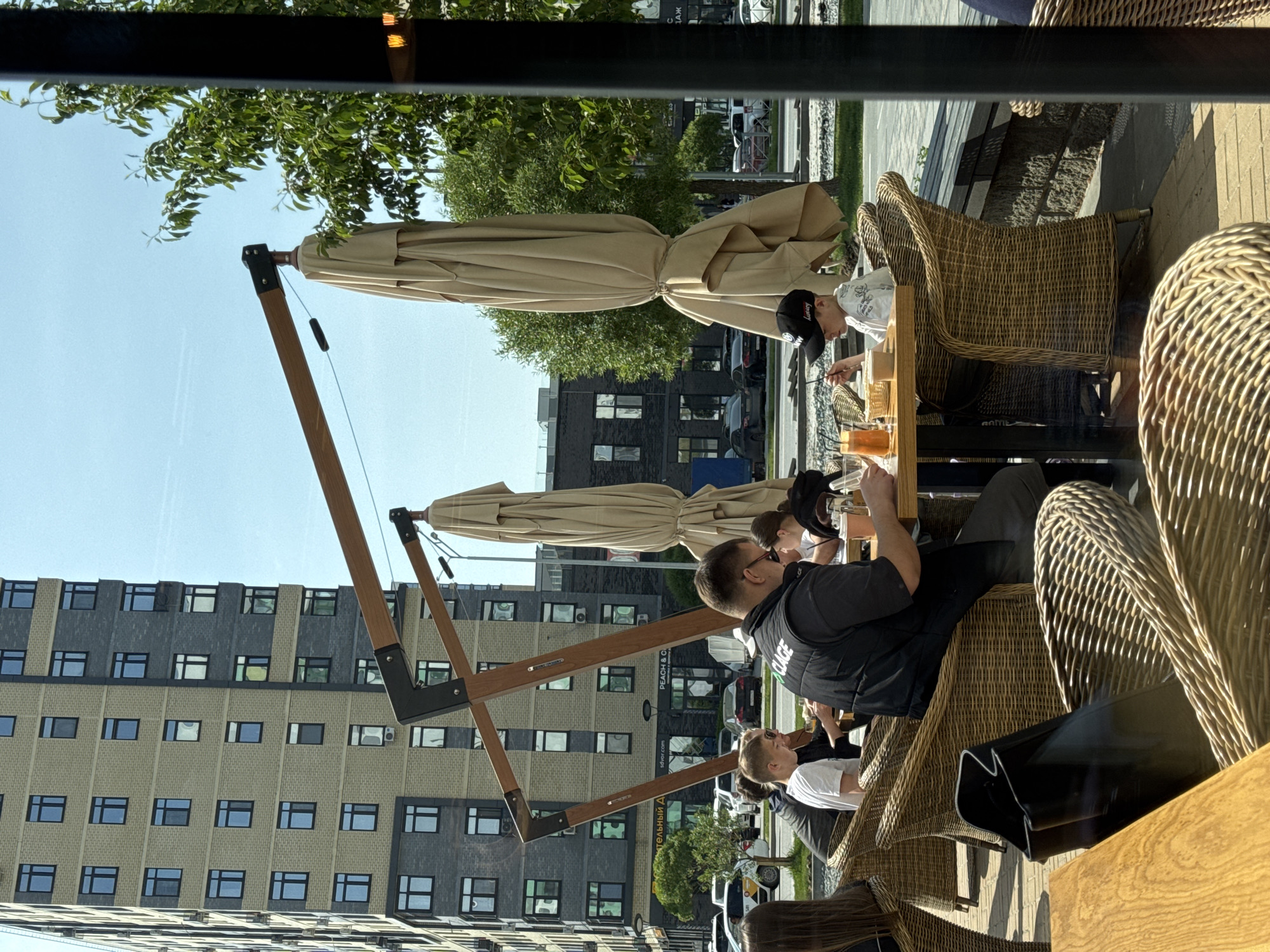 Captured through a window, the scene depicts an outdoor patio in Tyumen, Russia. Several people are seated at wicker chairs and tables under a large beige parasol. The diners appear to be engaged in conversation and enjoying their meals. In the background, a tall, modern apartment building dominates the skyline. The architectural style of the building and the overall ambiance suggest a contemporary urban setting.