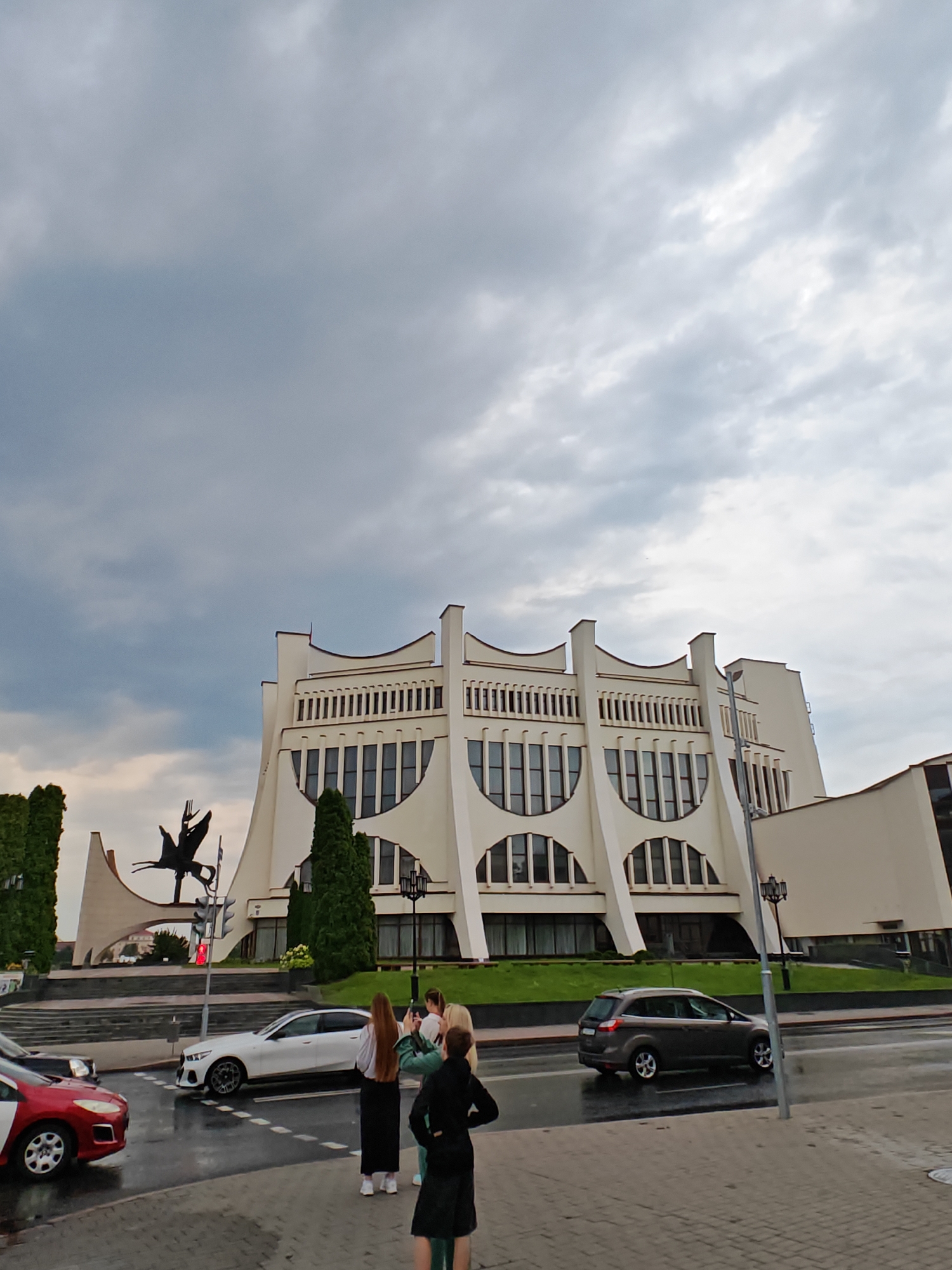 This is a wide-angle, eye-level photograph of a modernist building, likely the Hrodna Regional Drama Theatre, in Hrodna, Belarus. The building is a light-colored, multi-story structure with a distinctive facade featuring a series of arched windows and vertical decorative elements. In front of the building, to the left, stands a dark metal sculpture of a winged creature or figure. The sky is overcast with dramatic, grey clouds. In the foreground, a wet street with painted lane markings is visible, suggesting recent rain. Several cars are present: a red and white vehicle on the left, a white sedan turning left in the middle, and a dark gray SUV on the right. Four individuals are standing on the sidewalk on the right side of the frame, some appearing to be taking pictures with their phones. The overall impression is a somber, overcast day in an urban setting with prominent architecture.