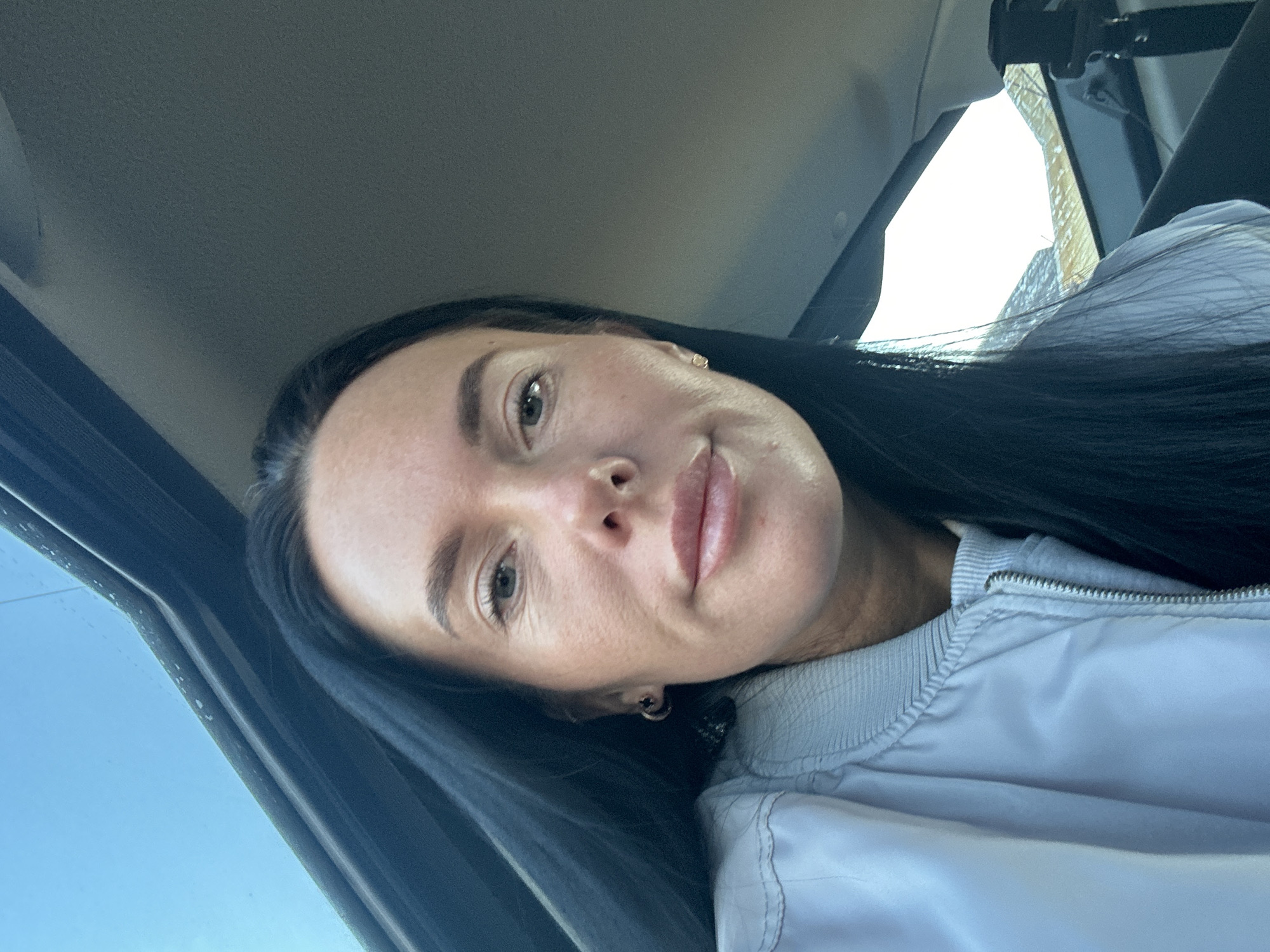 A woman with dark hair and light skin is inside a vehicle. The background shows parts of the vehicle's interior (roof, window frame) and a glimpse of a building outside, potentially indicating an urban setting. It is not possible to determine if the location is in Ольховка, Russia based solely on the image.