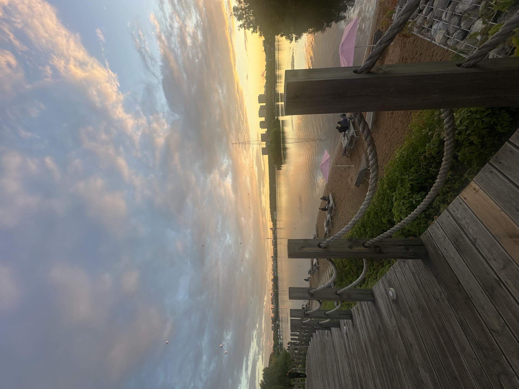 This image captures a serene waterside scene in Myakinino, Russia, likely during the late afternoon or early evening. The sky is a canvas of soft clouds tinged with the warm hues of sunset, reflecting on the calm water. A wooden boardwalk with rope railings leads the eye into the scene, suggesting a recreational area.
On the sandy beach beyond the boardwalk, several pink umbrellas are dotted, hinting at a place where people might relax. Some individuals are visible sitting on benches, enjoying the tranquil atmosphere. In the distance, buildings and a large electrical tower punctuate the horizon, providing a sense of urban proximity to this natural setting. The overall mood is peaceful and relaxing, amplified by the gentle light and calm waters. There is no visible text in the image.
