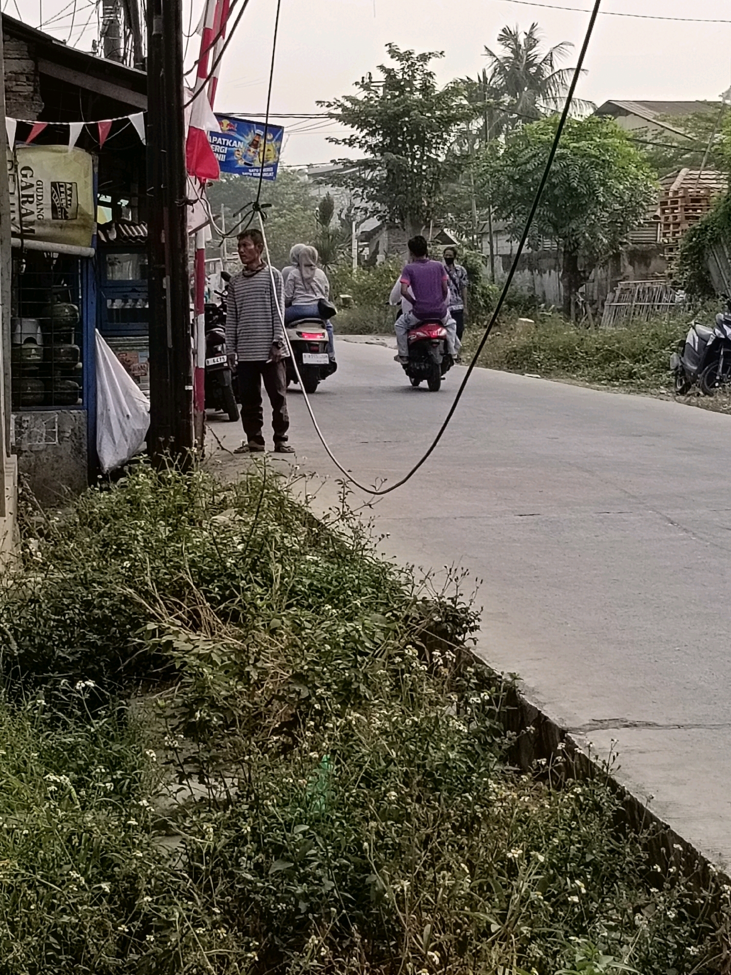The image depicts a street scene in Belimbing, Indonesia, likely during daylight hours with overcast skies. In the foreground, lush green vegetation, including small white flowers, lines a concrete ditch adjacent to a paved road. A man in a striped long-sleeved shirt and dark pants stands near a utility pole on the side of the road, holding what appears to be a cigarette. Behind him, a woman wearing a light-colored hijab sits on the back of a scooter, facing away from the camera. A second scooter, ridden by a person in a purple shirt, is visible further down the road, also moving away from the viewer. Another individual stands on the side of the road behind the purple-shirted rider.
To the left of the road, a small building with a partially brick facade is visible, adorned with red and white triangular pennants. A large yellow sign with black text, possibly advertising "GUDANG GARAM," is mounted on the building. Several green cylindrical objects, possibly gas tanks, are stacked in front of the building. A white tarp or fabric is also present.
The background shows trees, houses with simple architecture, and stacks of wood or building materials. Power lines crisscross the scene overhead. The overall impression is of a rural or semi-urban environment in Indonesia.