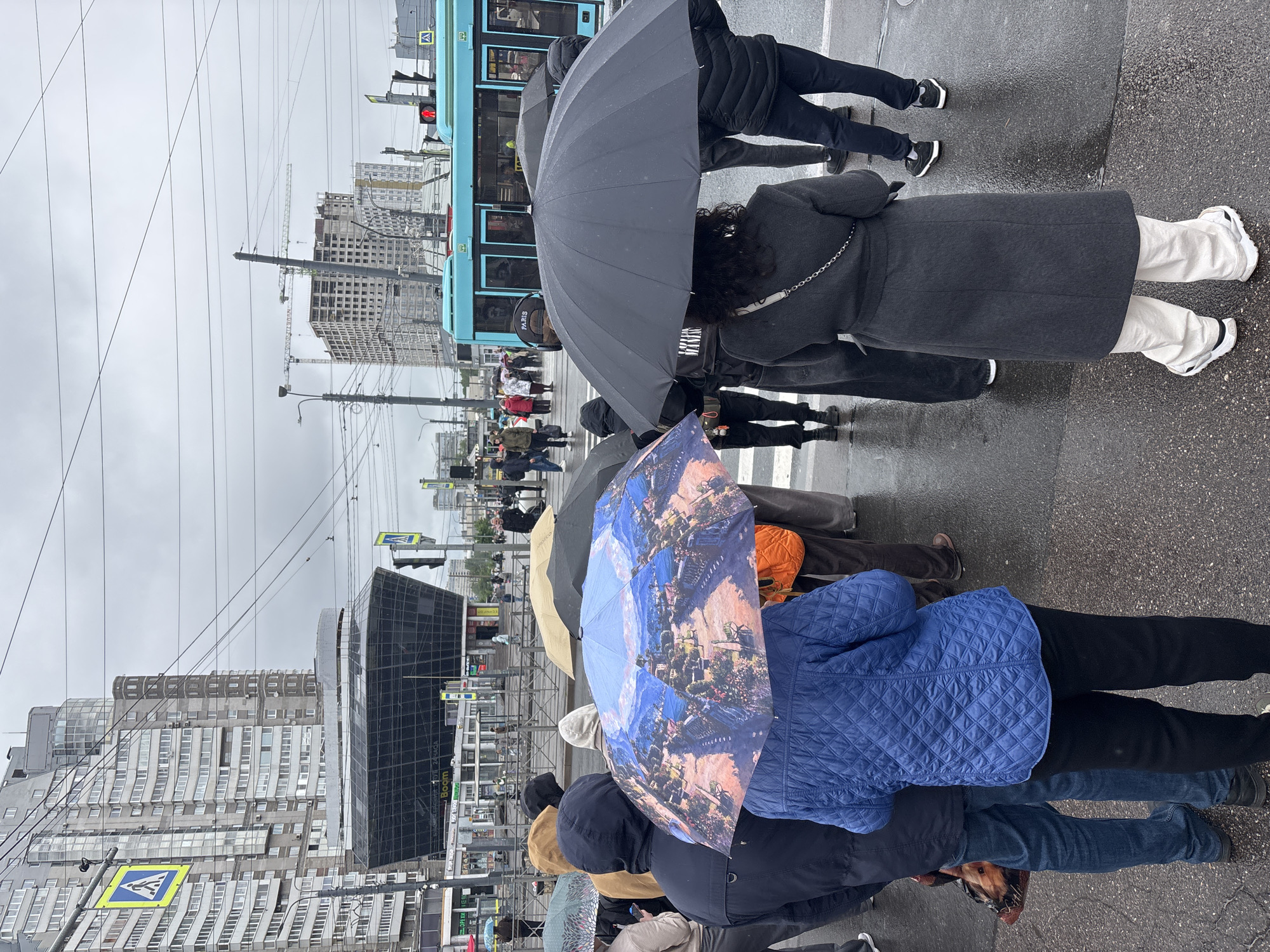 The image shows a group of people waiting at a crosswalk in Saint Petersburg, Russia, on a rainy day. Most are holding umbrellas, including one with a blue, pink, and white design. A blue tram is stopped nearby. The environment includes multiple buildings of varying heights and architectural styles, construction cranes, power lines, streetlights, and street signs. The ground is wet, indicating recent rainfall. The overall scene conveys a typical urban situation in inclement weather.