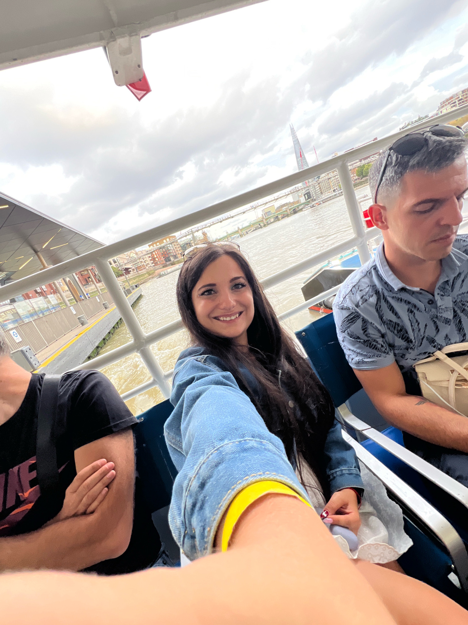 The image captures a selfie on a ferry in London, United Kingdom. The foreground features three individuals, with a woman in a denim jacket smiling at the camera. To her right, a man with sunglasses perched on his head looks downwards, and on the left, a person in a black t-shirt is partially visible with arms crossed. The ferry appears to be moving along a body of water, likely the River Thames, with a cityscape in the background. Notable structures visible in the distance include The Shard, a prominent skyscraper. The environment suggests an overcast day with a cloudy sky. The ferry has a white railing and dark blue seats.