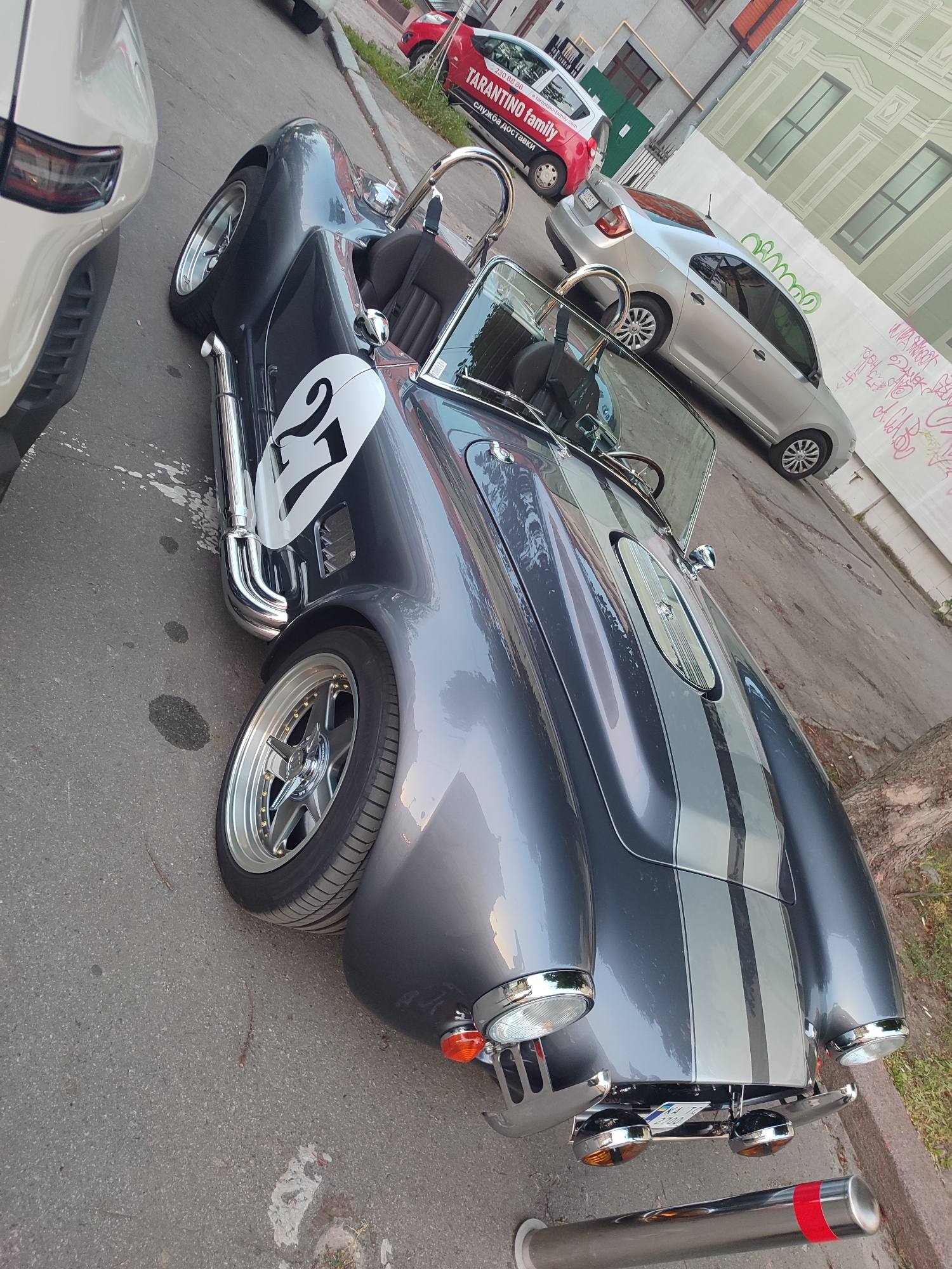 This media file features a low-angle, eye-level shot of a dark grey classic sports car, likely a replica of a Shelby Cobra, parked on an asphalt street. The car has a distinctive silver racing stripe down the center and the number "27" in a white circle on its front fender. Chrome accents, including the side pipes, roll bar, and windshield surround, gleam in what appears to be natural daylight. The car is positioned in a street scene with other vehicles and buildings visible in the background. To the left, part of a lighter-colored vehicle, possibly a modern SUV or sedan, is in the frame. Further back, a red compact car with "Tarantino family" branding and Cyrillic text is parked alongside a silver sedan. The buildings in the background appear to be a mix of older, multi-story structures with windows and a lighter-colored wall covered in graffiti. The ground shows some oil stains. The overall scene suggests an urban environment during daylight hours.
