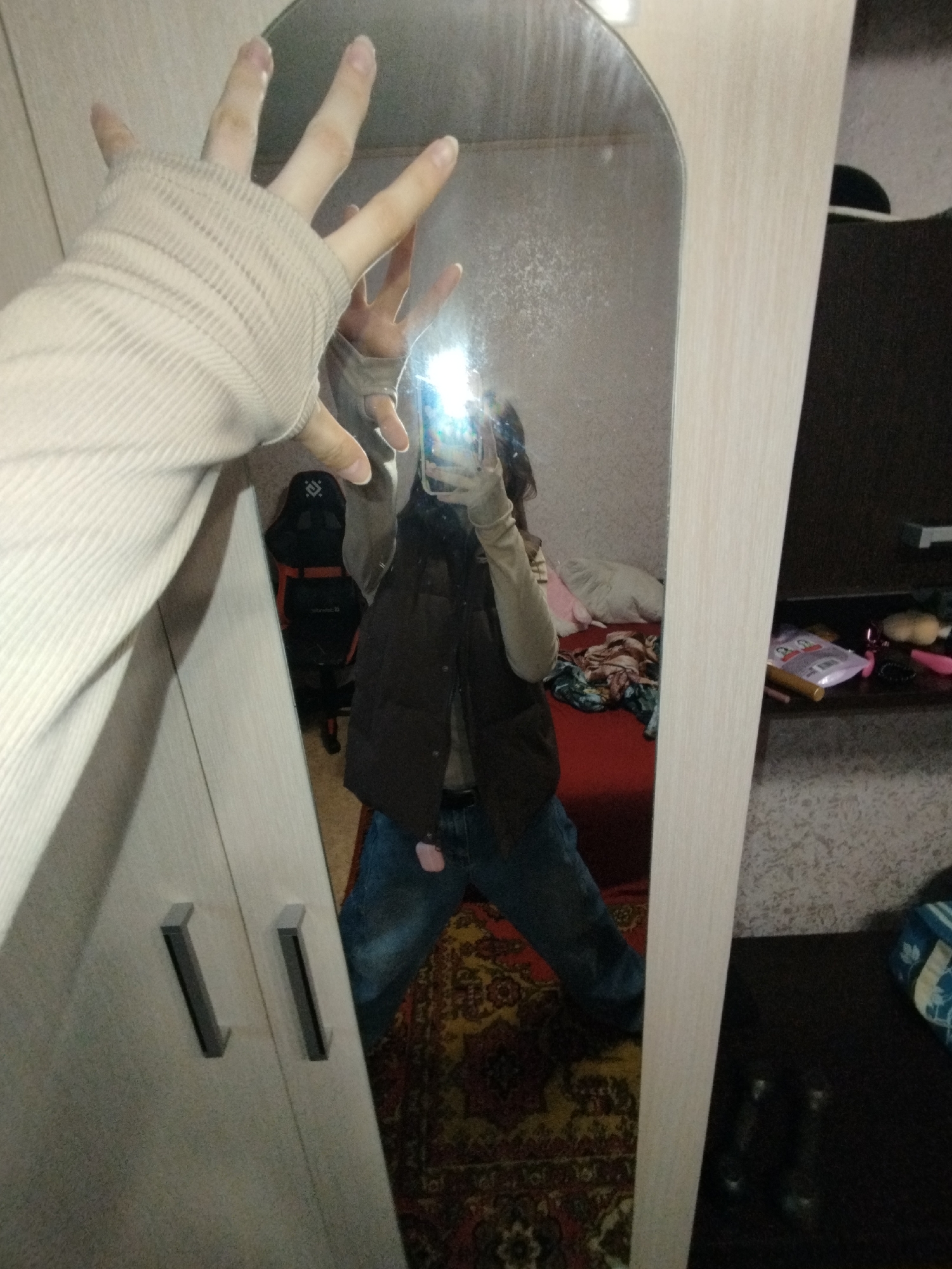 A person is taking a photo of themself in a mirror. The person is wearing a light beige long-sleeved shirt, a brown vest, and blue jeans. The person's left hand is extended toward the mirror, fingers spread. A smartphone is visible in the person's right hand, the screen illuminated.
The mirror is part of a wardrobe or cabinet with long, vertical metal handles. The background reflects the person's bedroom, which includes a bed with red bedding and scattered clothes, a black gaming chair, and a dark wooden desk with various items.
The floor is covered with a patterned carpet. The location is likely a private residence, and based on the presence of Cyrillic text on items in the reflection, the location is likely in Russia. Based on the context, this may be in Saratov.