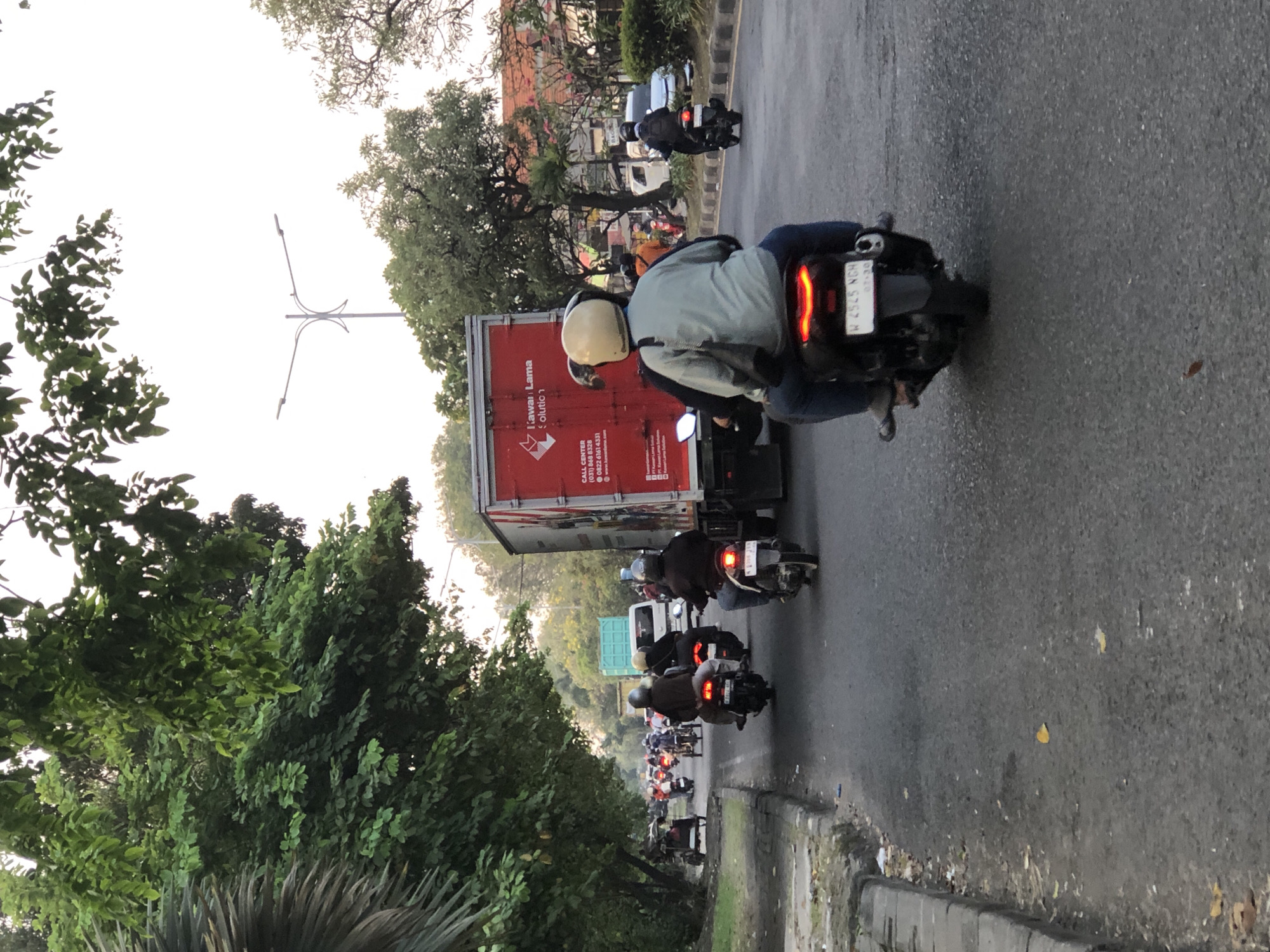 In Sumorame, Indonesia, a busy street scene unfolds with numerous motorcycles and a large truck. The image captures traffic on an asphalt road lined with trees and a sidewalk. In the foreground, two people are riding a motorcycle. The rider wears a light blue jacket and dark pants, while the passenger is seated behind. The motorcycle has a prominent red taillight and a license plate displaying "W 4545 NGH". Further down the road, other motorcycles and their riders are visible, many wearing helmets and casual clothing. A large red truck with "Tianwan Lama" written on its side is prominent on the right side of the road, seemingly stopped or moving slowly. A teal truck is also present in the distance. The environment appears to be a typical urban or suburban setting with lush green trees providing shade. The sky is overcast, suggesting a potentially hazy or diffused light. The overall impression is one of a bustling day in Sumorame, with motorcycles being a primary mode of transportation.