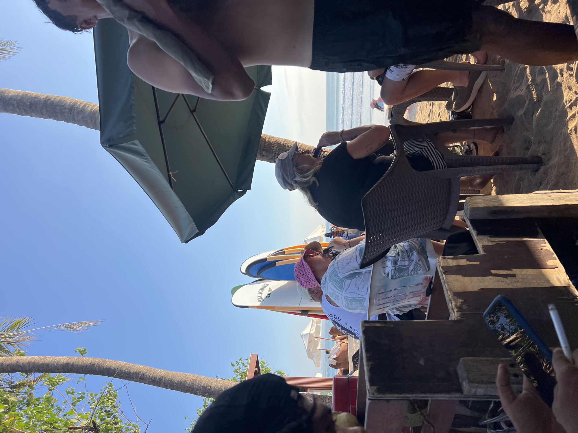 The image depicts a beach scene in Kuta, Indonesia, with several people gathered at tables set on the sand. In the foreground, a person holds a smartphone and a cigarette. Behind them, a group of individuals are seated at tables, with surfboards standing upright in the background. One person wears a pink bucket hat, another a grey baseball cap, and a third has blonde hair. Palm trees are visible, casting shadows on the sand. The ocean and a clear blue sky form the backdrop. Beach umbrellas and lounge chairs are also present further down the beach.