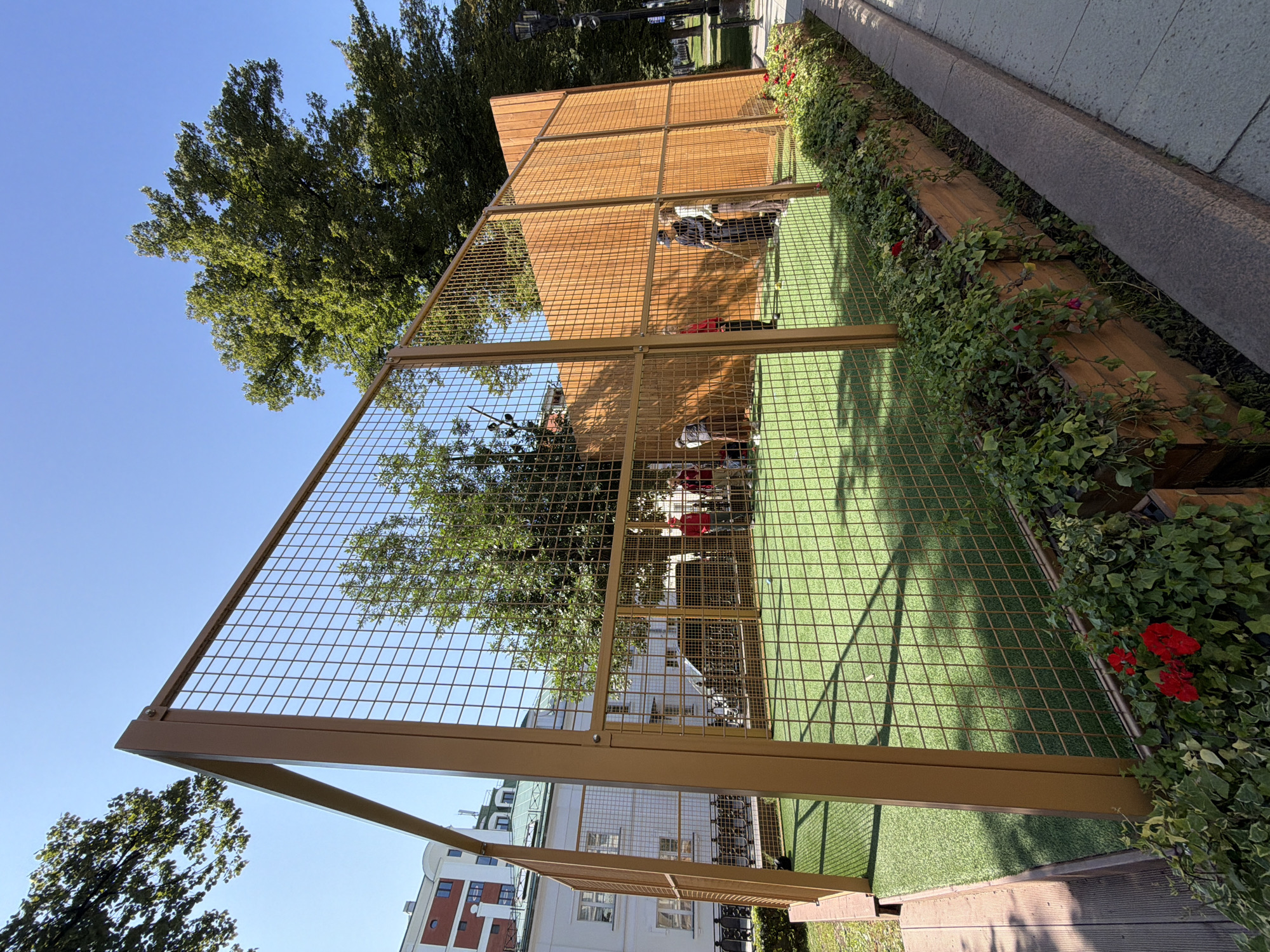 The image depicts an outdoor court with an artificial turf surface, enclosed by a metal mesh fence and a wooden back wall. Several people are present, engaged in an activity resembling a sport. In the background, there are buildings, trees, and a clear blue sky. The court is lined with planter boxes containing greenery and red flowers. This scene is located in Moscow, Russia, specifically on Bolotnaya Square.