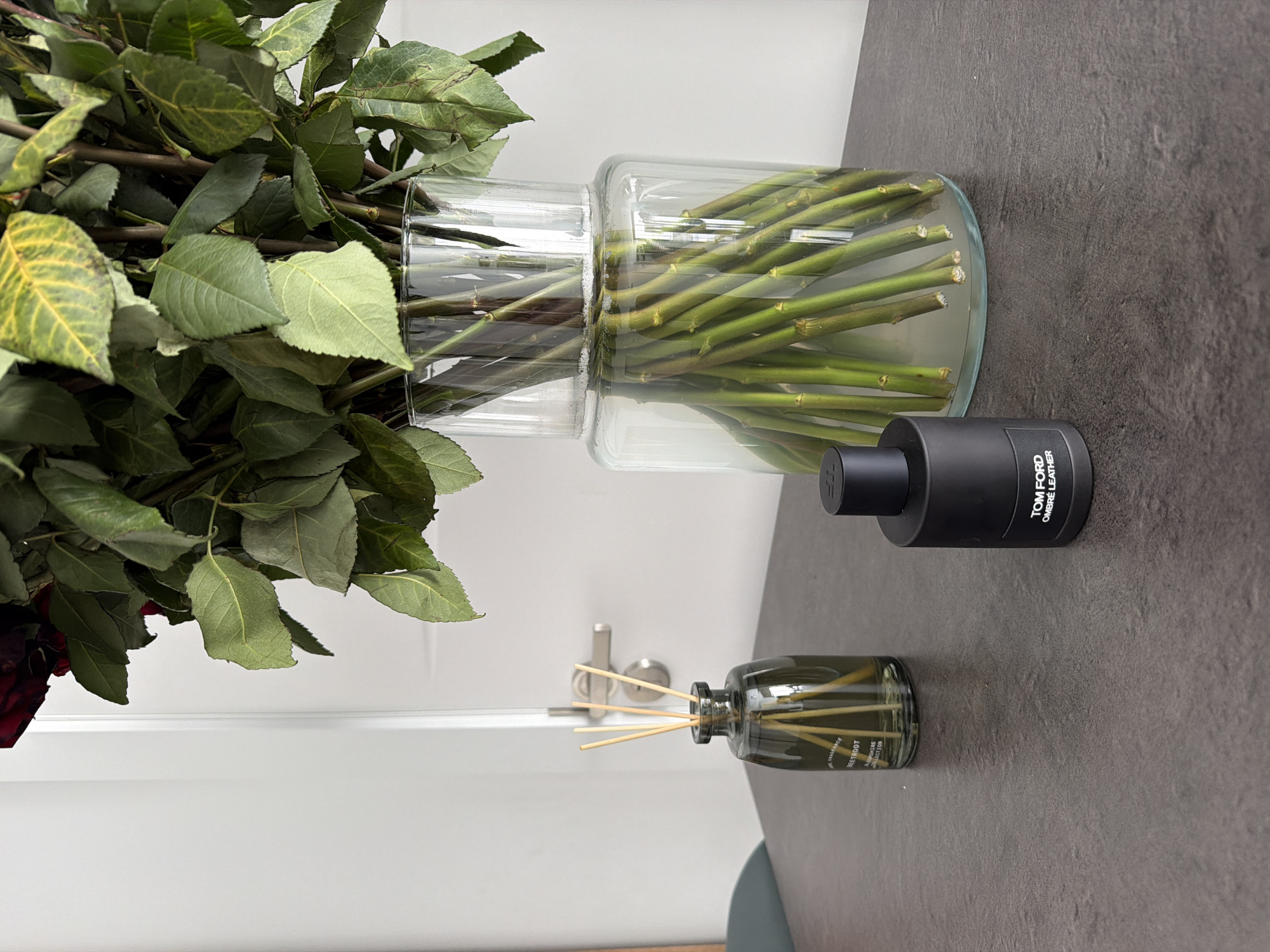 A dark gray textured surface, likely a table, occupies the foreground of the image. On this surface, there are three objects. To the left, a dark green glass diffuser bottle with several reeds is positioned. To the right of the diffuser is a black cylindrical perfume bottle with a black cap, labeled "TOM FORD OMBRÉ LEATHER". Behind these objects, a large, clear glass vase filled with a bouquet of red roses and green foliage is visible. The background consists of a white door and door handle, indicating an indoor setting. No people are present. The lighting suggests a well-lit indoor space. The scene does not provide any information to determine its location in Finestrat, Spain.