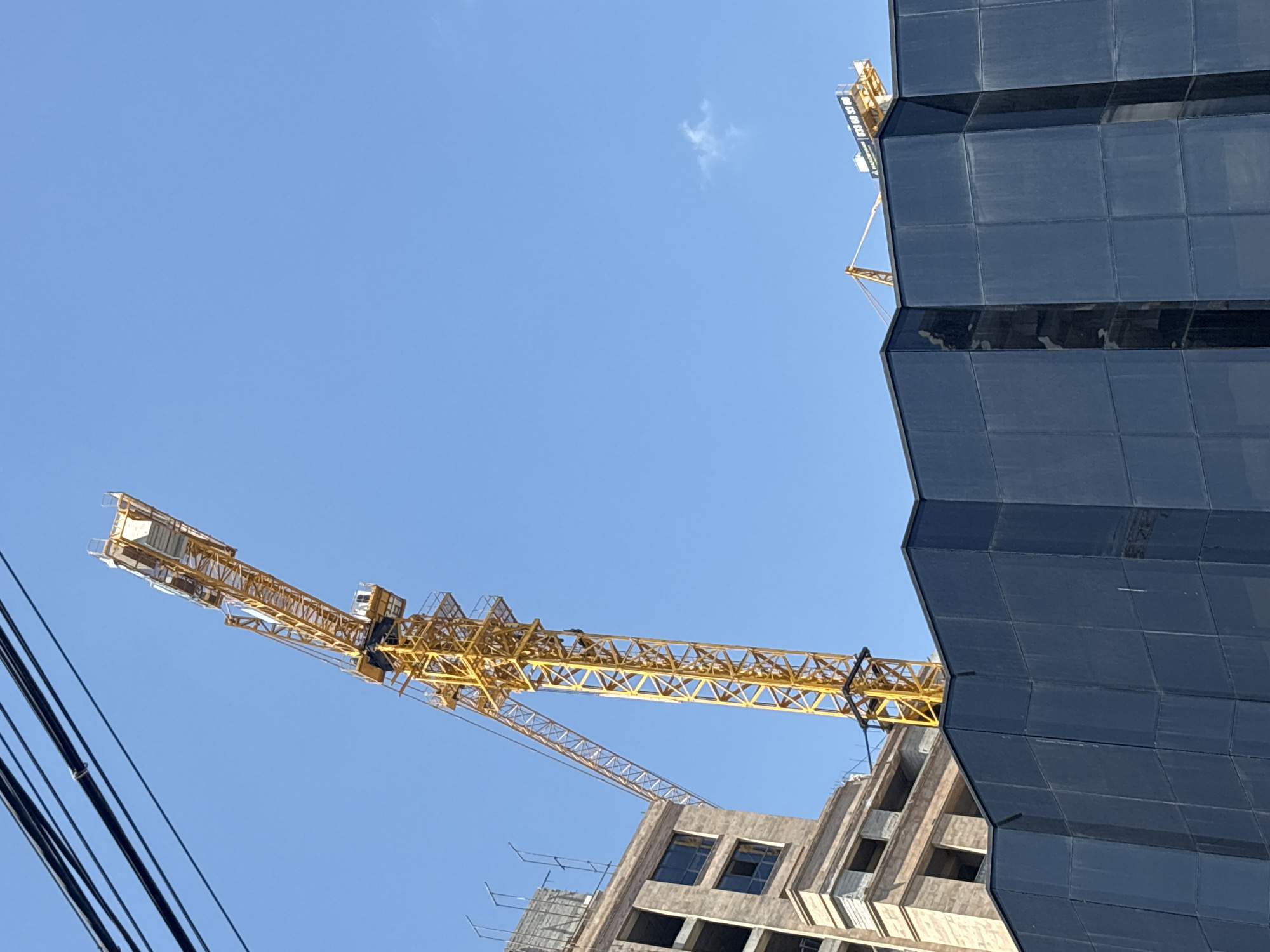 The image depicts a construction site in Yerevan, Armenia. A yellow construction crane, composed of a lattice structure, extends diagonally across the frame against a clear blue sky. Parts of the crane are positioned above a building under construction. A modern skyscraper is visible on the right side of the frame, its exterior covered in dark, protective sheeting or cladding. The lower portion of the building under construction reveals an unfinished concrete facade with openings for windows. Electrical wires are visible in the lower left corner. There are no people visible in the frame.