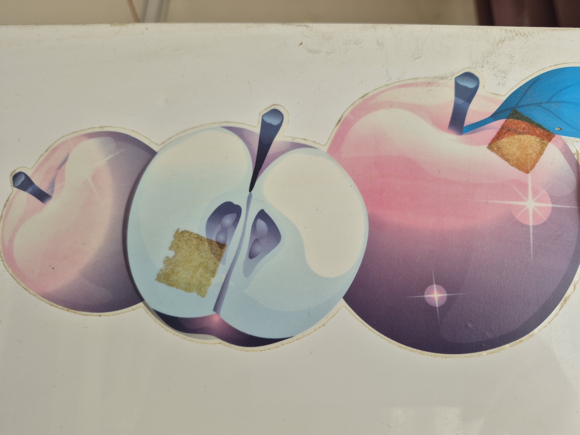 The image features a close-up of a decorative graphic on a white, possibly laminated or plastic surface. Three stylized apples are depicted with a glossy, semi-transparent appearance. The leftmost apple is partially visible, featuring a pinkish-purple color gradient and a dark blue stem. The central apple is shown cut in half, revealing light blue inner flesh with darker blue seed-like structures and a dark blue stem. A small, irregularly shaped piece of light brown residue, possibly adhesive or a tear, mars the surface of its left half. The rightmost apple is fully visible, large and predominantly purple with pink highlights, a dark blue stem, and an attached light blue leaf. This apple also has a rectangular piece of light brown residue near its stem and several white starbursts indicating sparkle. The apples are outlined on the white background, which shows minor dirt and wear, particularly along the edges of the apple shapes. No activity or event is taking place, and no text is visible. While the location context provided is Yerevan, Armenia, there are no specific visual cues within the image itself to confirm this setting.