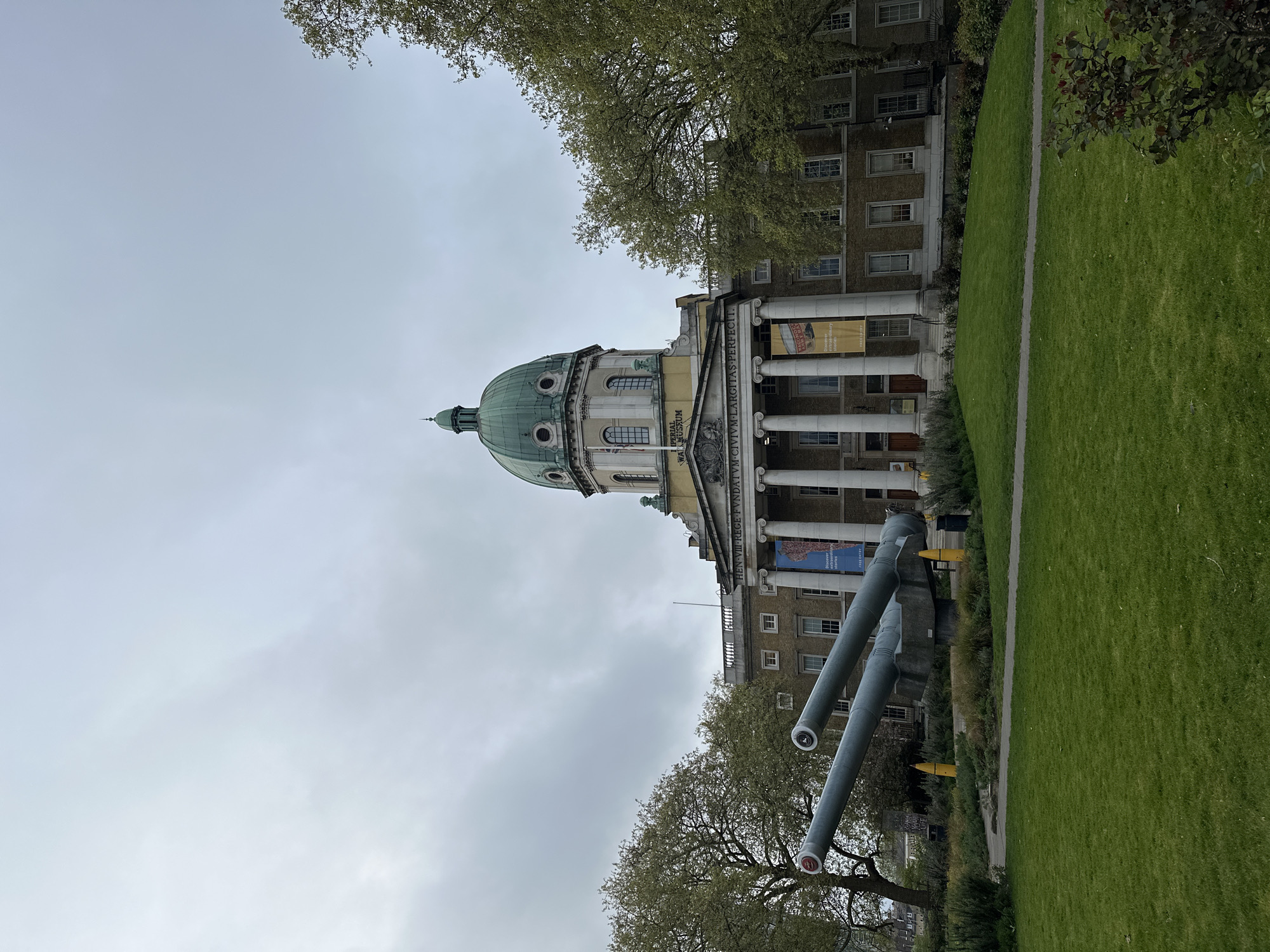 The Imperial War Museum in London, United Kingdom, is seen from a low angle on an overcast day. The building has a large green dome, classical columns, and is fronted by two large naval guns pointing towards the sky. Adjacent to the museum is a grassy area with trees.