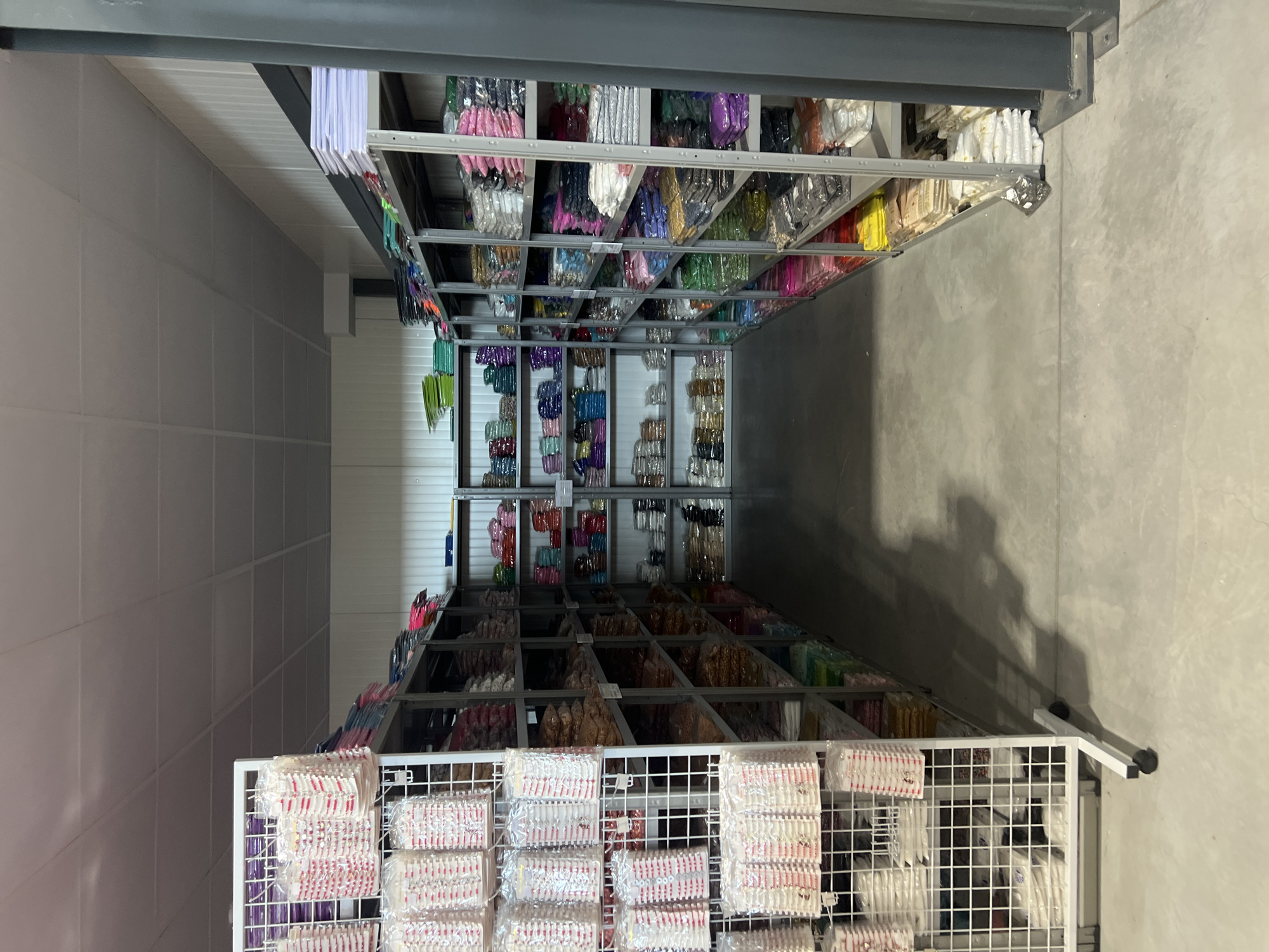 The image shows an interior view of a storage facility, likely a warehouse, in Burgas, Bulgaria. Shelving units laden with packaged goods, possibly small items or merchandise, line the walls. The items appear to be sorted by color and type. The flooring is concrete, and the walls are composed of metal panels. The lighting is provided by overhead fixtures. A wire rack with additional packaged goods is visible in the foreground. The setting suggests a commercial or industrial storage space.