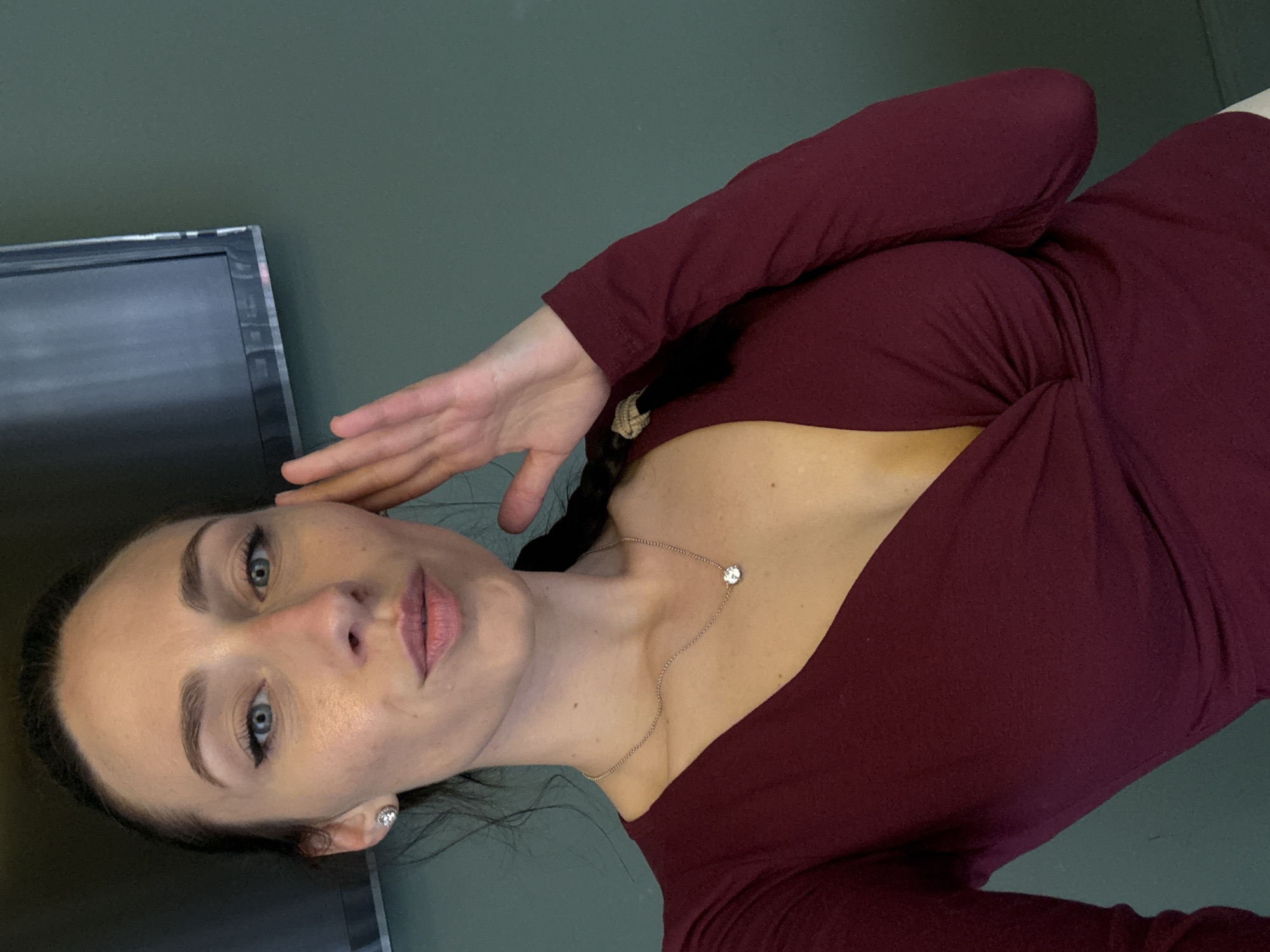 A woman with fair skin, blue eyes, and dark hair braided at the back poses in a portrait. She wears a long-sleeved maroon dress and a delicate necklace with a small circular pendant. Her left hand is raised near her face. In the background, a corner of a television screen is visible against a green wall. The location is not identifiable as Saint Petersburg, Russia, without additional contextual clues.
