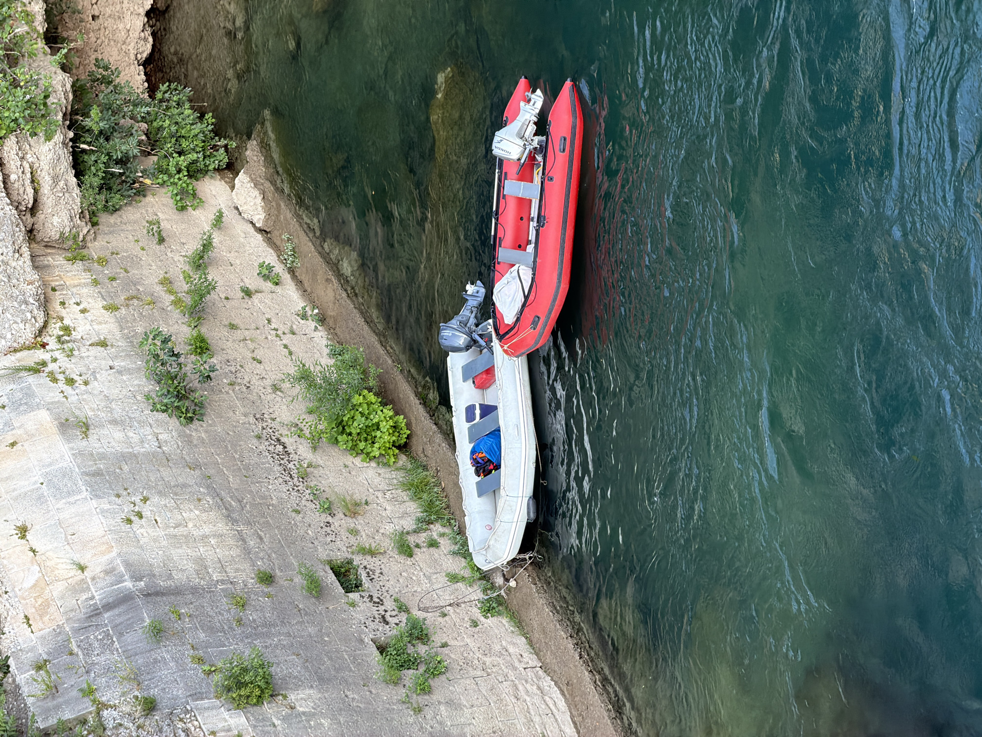 Two inflatable boats are moored to a stone wall on the bank of a body of water, likely the Neretva River, in Mostar, Bosnia and Herzegovina. The boats are an off-white dinghy with a blue and red bag on board and a bright red inflatable boat with two outboard motors. The stone wall is weathered and shows signs of vegetation growth. The water is a deep green color.