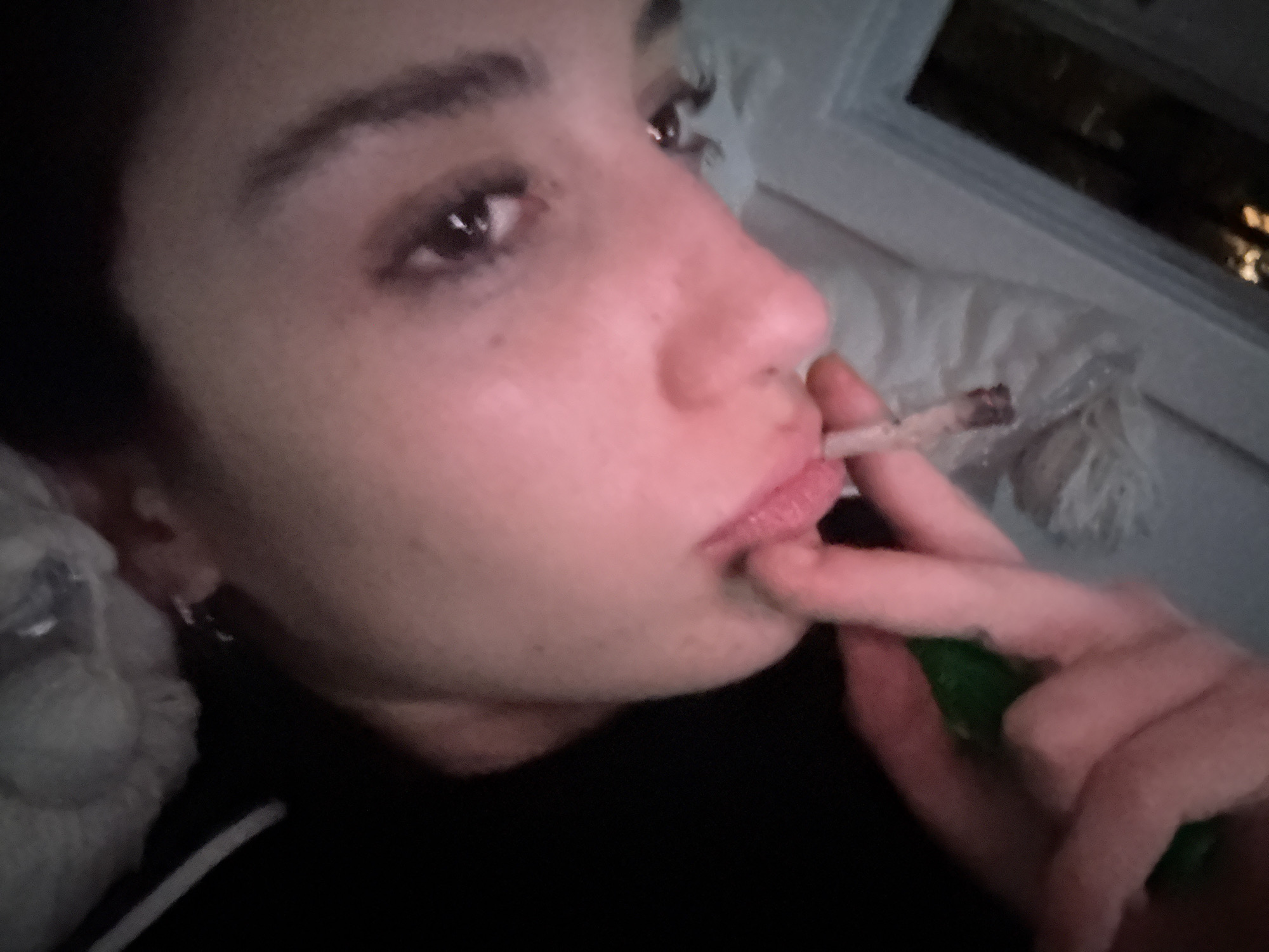 A close-up selfie captures a woman with light skin, dark hair, and dark eyes. She is holding a lit cigarette or joint between her lips and fingers. She wears an earring in her left ear. The background includes a pillow, a window, and an indistinct view outside the window at night. There is no information in the image that indicates the location is Buenos Aires, Argentina.