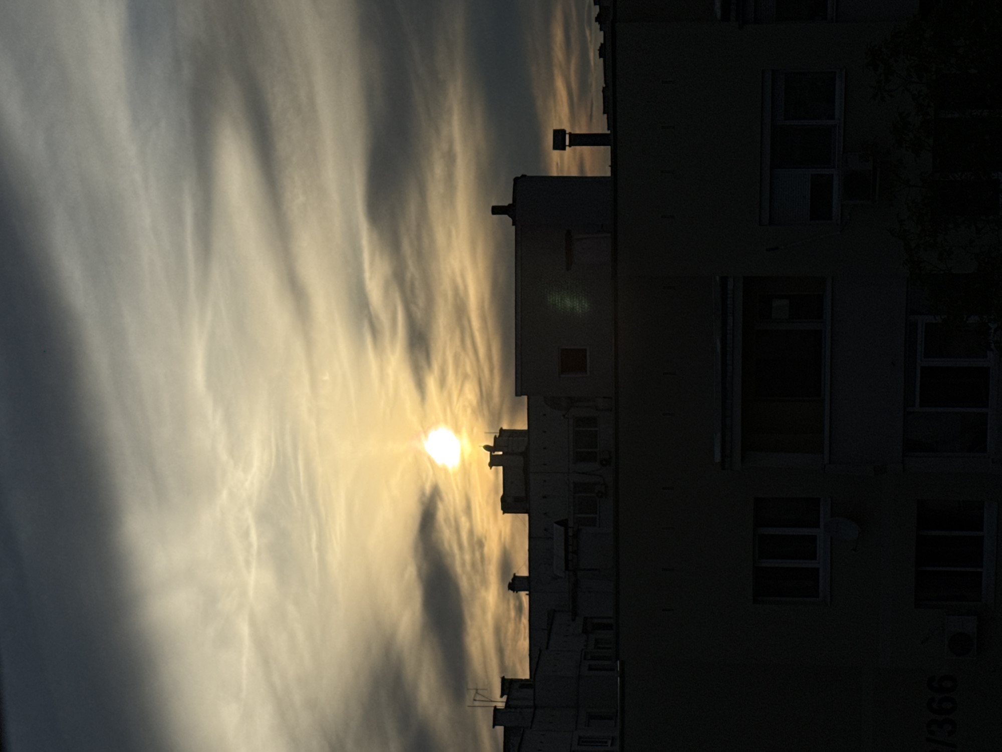 The image shows a building in the city of Pleven, Bulgaria, likely an apartment building, viewed against a backdrop of the sky. The sky is partially obscured by clouds, with the sun shining through, creating a bright glow. The building appears to be several stories tall, with windows and some architectural details visible, despite being in shadow. A number is visible at the bottom right which appears to be 2/366. Trees are visible to the top right of the building. There is no information about any people, actions, or interactions.