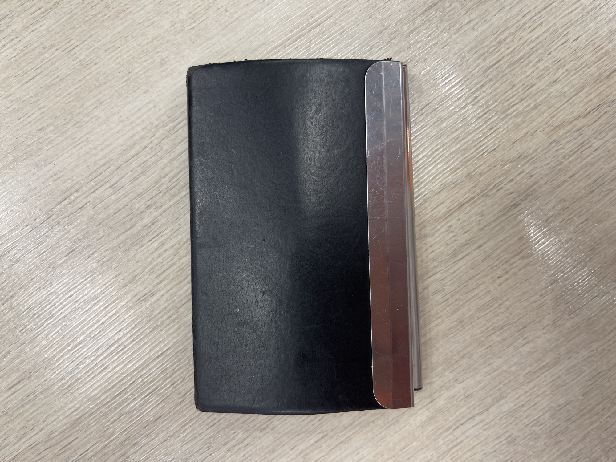 A black business card holder with a metallic trim rests on a light-colored wooden surface. The surface has a visible wood grain pattern. The business card holder is oriented horizontally, with its length running from left to right. The metallic trim is located along the bottom edge of the card holder. No people are visible in the image, and no specific location in Moscow, Russia can be identified from the provided image.