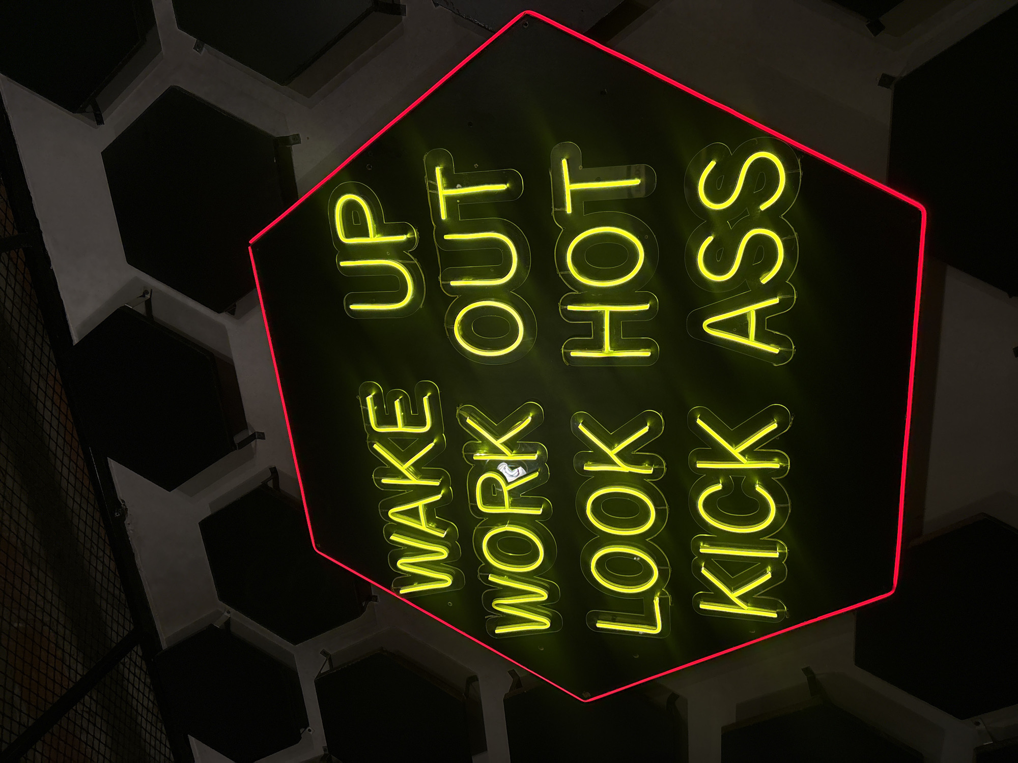 A low-angle shot shows a neon sign on a black background, framed by a red neon outline in a hexagon shape. The text "WAKE UP, WORK OUT, LOOK HOT, KICK ASS" is written vertically in bright yellow neon lights. The background appears to be a ceiling with hexagonal panels and some metal mesh. The location, San Bernardino Tlaxcalancingo, Mexico, cannot be visually confirmed in the image.
