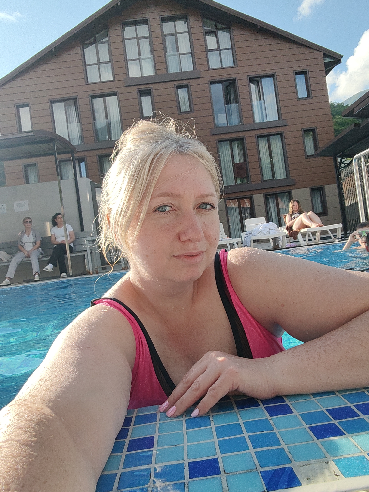 A blonde woman with freckles, wearing a pink and black swimsuit, is taking a selfie in a swimming pool. She is leaning on the edge of the pool, which is tiled with blue and white squares. In the background, a multi-story brown building with many windows is visible, possibly a hotel. Several other people are in the background, some sitting on benches, others on lounge chairs by the pool. One person appears to be a child in the water. The environment suggests a sunny day, with some clouds in the sky and a mountainous backdrop. The location is identified as Krasnaya Polyana, Russia, a resort town known for its ski slopes and mountain views.