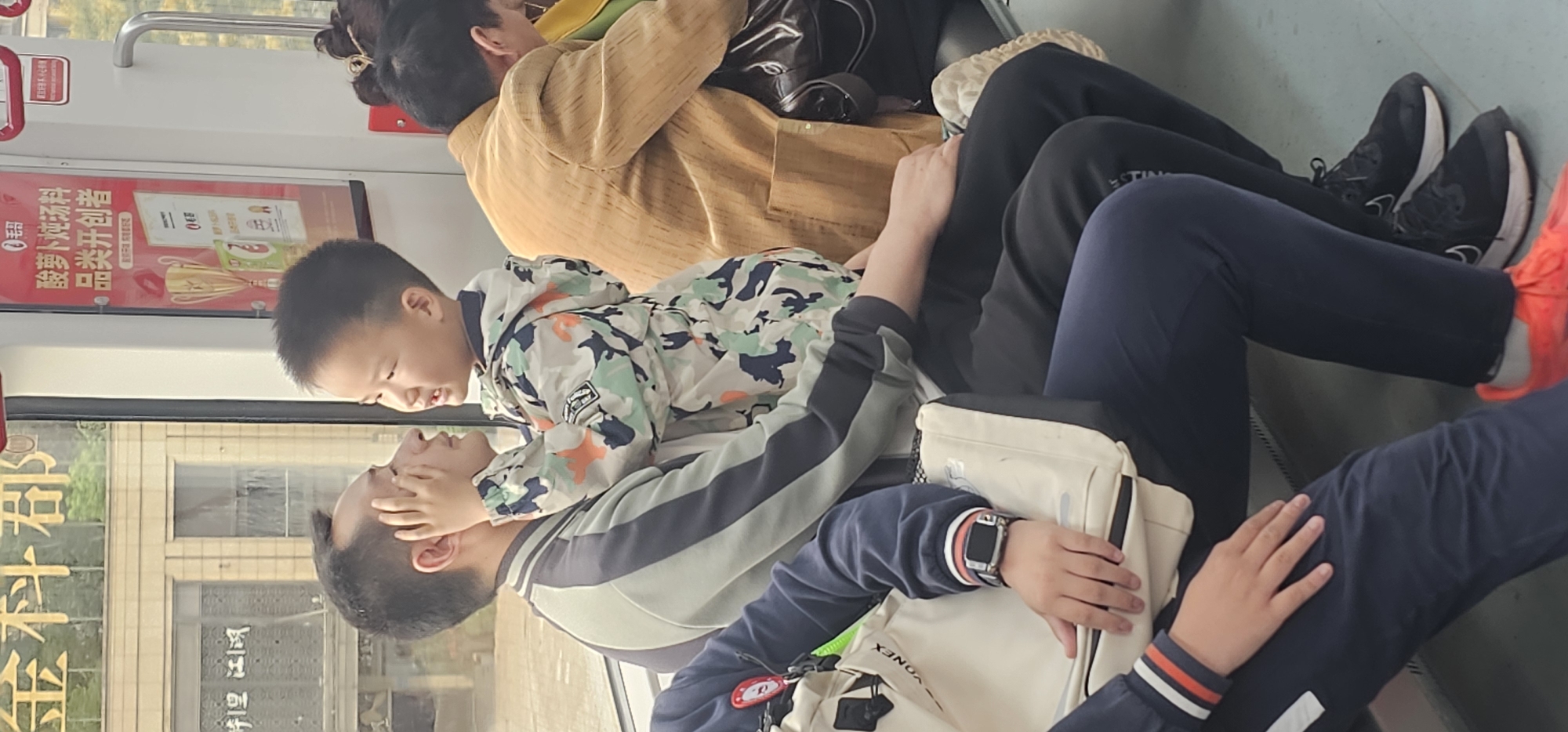The image captures a scene on what appears to be public transportation, possibly a subway train, in Chongqing High-tech Zone, China. Three individuals are visible. At the top of the frame, an adult with dark hair and wearing a light-brown jacket is seated and leaning slightly, with a dark bag nearby. Below, a child in a camouflage jacket is interacting with an adult who is wearing a jacket with white, gray, and blue accents. The child's hands are near the adult's face. The adult is wearing a watch. Another individual is seated to the right, wearing dark pants and bright orange/red shoes. An advertisement is visible above the child and adult. Outside the window, buildings and a sign with gold lettering are visible. The scene appears to be candid, capturing an ordinary moment.