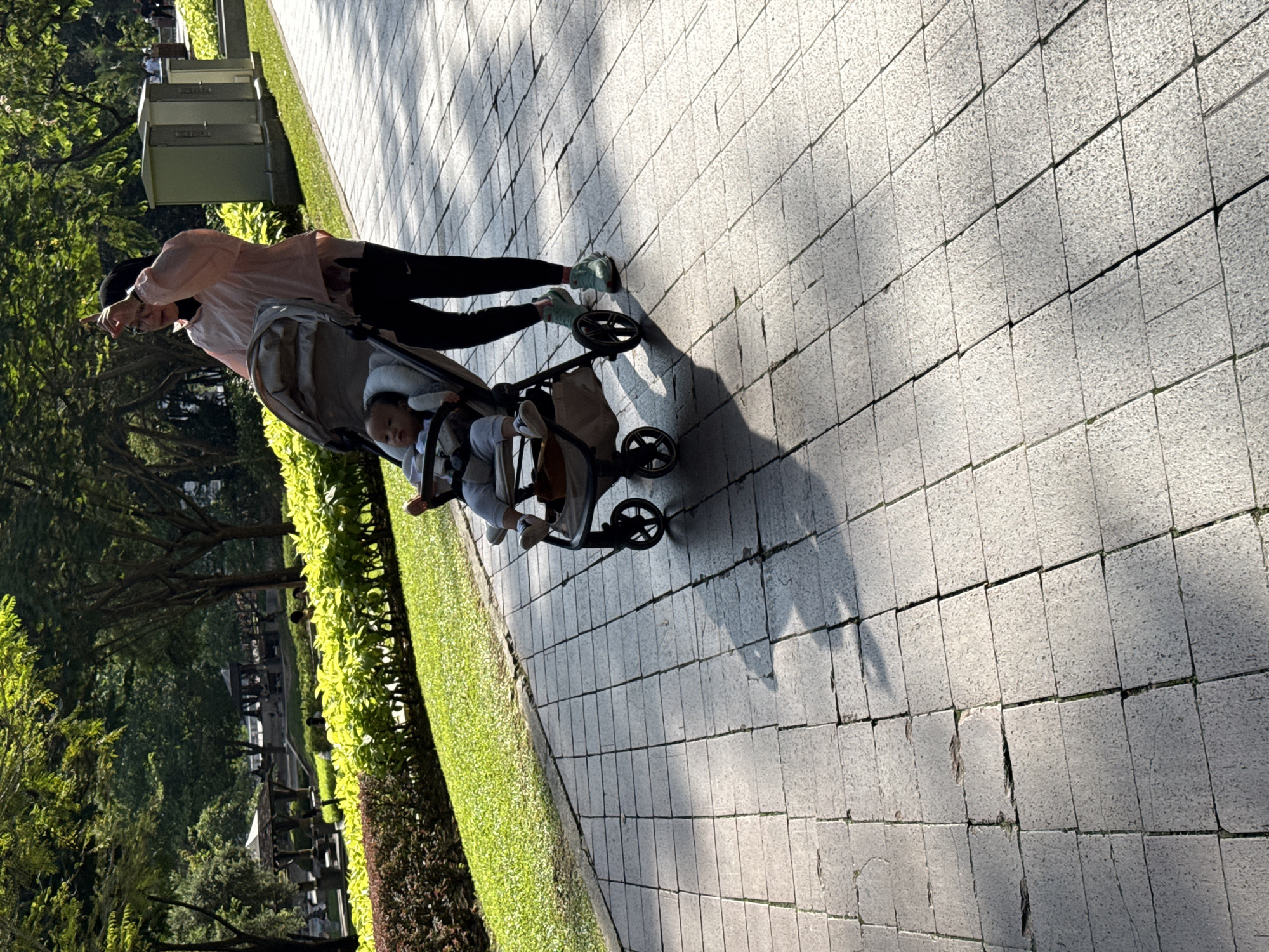 A woman in a hijab and light pink shirt pushes a stroller on a paved walkway in Kuala Lumpur, Malaysia. The stroller contains a baby. The walkway is bordered by a green lawn and trees. The walkway consists of rectangular gray pavers. The lighting suggests a sunny day. The photograph is taken at an angle.