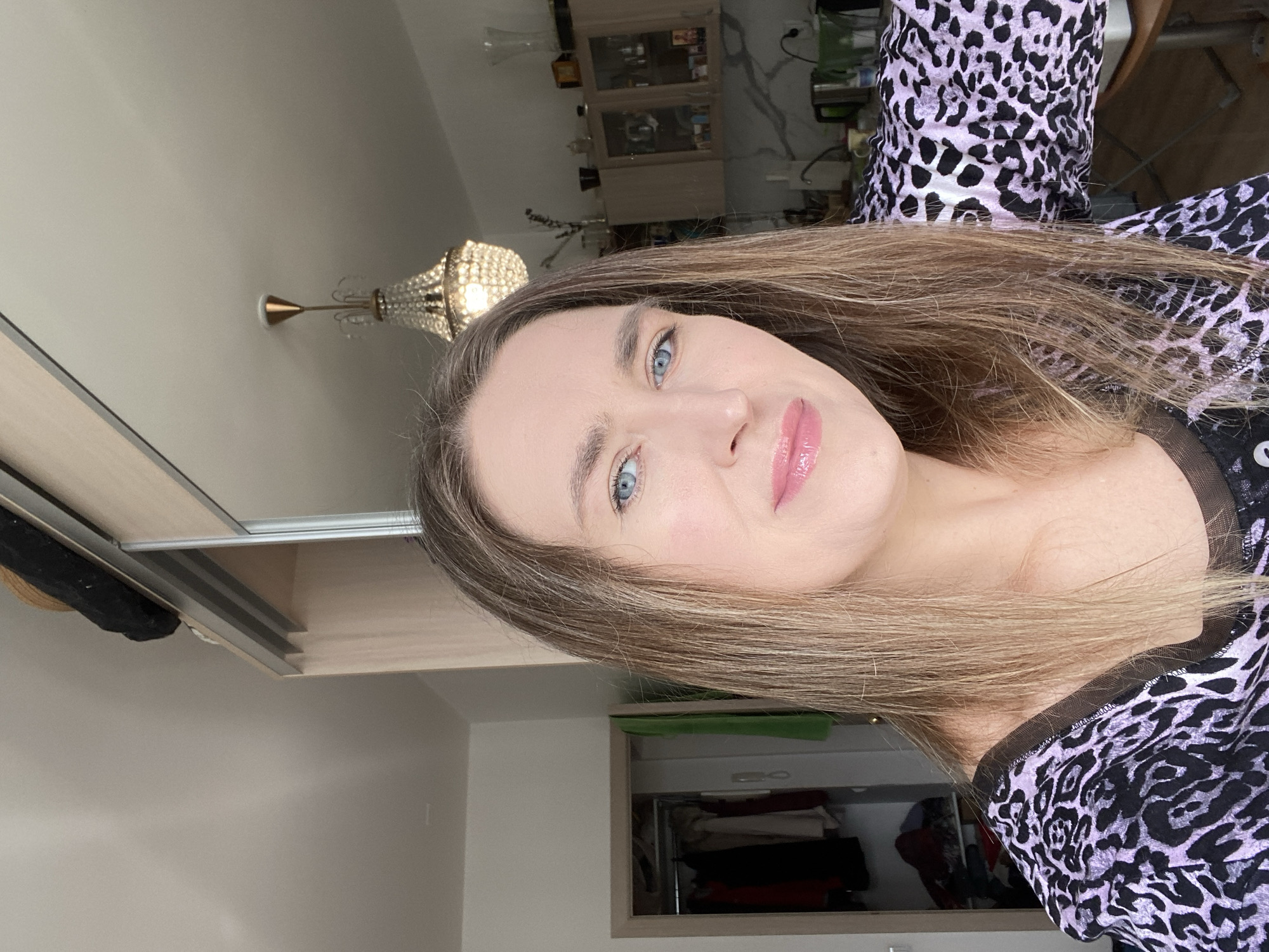 A woman with long brown hair and blue eyes is posing for a selfie indoors. She is wearing a purple and black leopard print top with a sheer black neckline. In the background, there is a beige wardrobe, a chandelier, and a display cabinet. The room appears to be in a residential setting. There is no clear indication of Yekaterinburg, Russia.