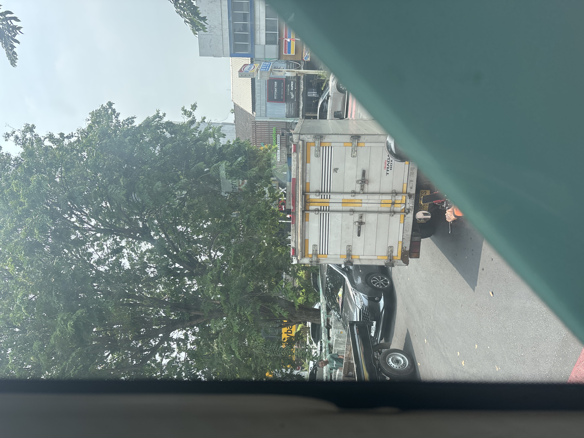 The image depicts a street scene in North Jakarta, Indonesia, likely during daylight hours with hazy skies. In the foreground, a portion of a light blue car's windshield and body is visible on the right. To the left, a black truck bed with a silver rim is partially seen.
Dominating the middle ground is a white box truck with yellow trim and silver latches. Its side features the text "ISUZU TRAGA BOX". Behind this truck, a dark gray sedan is stopped in traffic. A man wearing a green t-shirt stands on the left side of the road, near a low concrete barrier.
Further back, lush green trees with dense foliage line the street. Buildings with various signage are visible in the background, including a yellow sign with the text "yba" and another building with signs like "INFOBEAUTY", "CAR WASH", and "RUPEES". There is also a red and white striped crosswalk visible on the road.
The overall impression is a busy urban environment with moderate traffic and commercial activity. The presence of the box truck and sedan suggests transportation and commerce. The trees indicate an area with some greenery within the city.