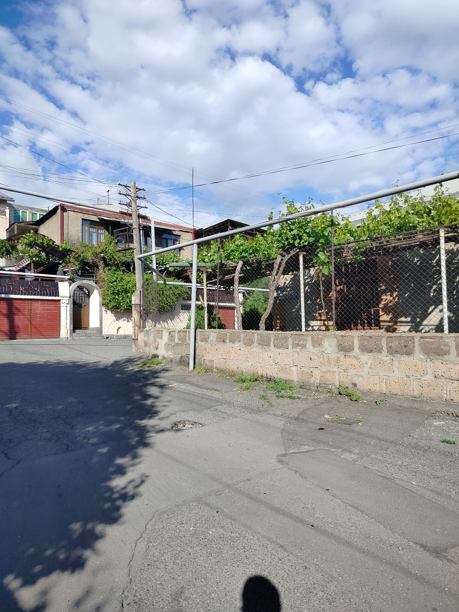 The image shows a residential street in Yerevan, Armenia. The street is paved with asphalt and shows signs of wear and tear. A low brick wall runs along the side of the street, topped with a metal fence covered in grapevines. Behind the fence is a building with red doors and a gated entrance. Utility poles and wires are visible above the buildings, set against a cloudy blue sky. The shadow of the photographer is visible at the bottom of the image.