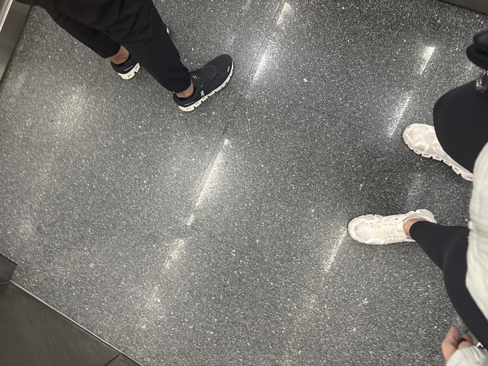 An overhead shot captures the lower legs and feet of two individuals standing on a speckled gray floor in San Bernardino Tlaxcalancingo, Mexico. The floor appears to be a stone material like granite or terrazzo, with scattered light and dark specks. Distinct, elongated streaks of bright white light are reflected on the floor, suggesting overhead illumination.
On the left, a person wears black pants and black athletic shoes with prominent white soles and the "On" brand logo visible on the side. On the right, another person wears black leggings and white athletic shoes, also featuring the "On" logo. A light-colored garment, possibly a jacket or hoodie, is partially visible on the upper right side of this individual. A metallic edge or rail runs along the bottom left corner of the frame. The individuals appear to be standing or moving within an indoor or covered public space. No specific activity or emotion is discernible beyond their presence in the scene.