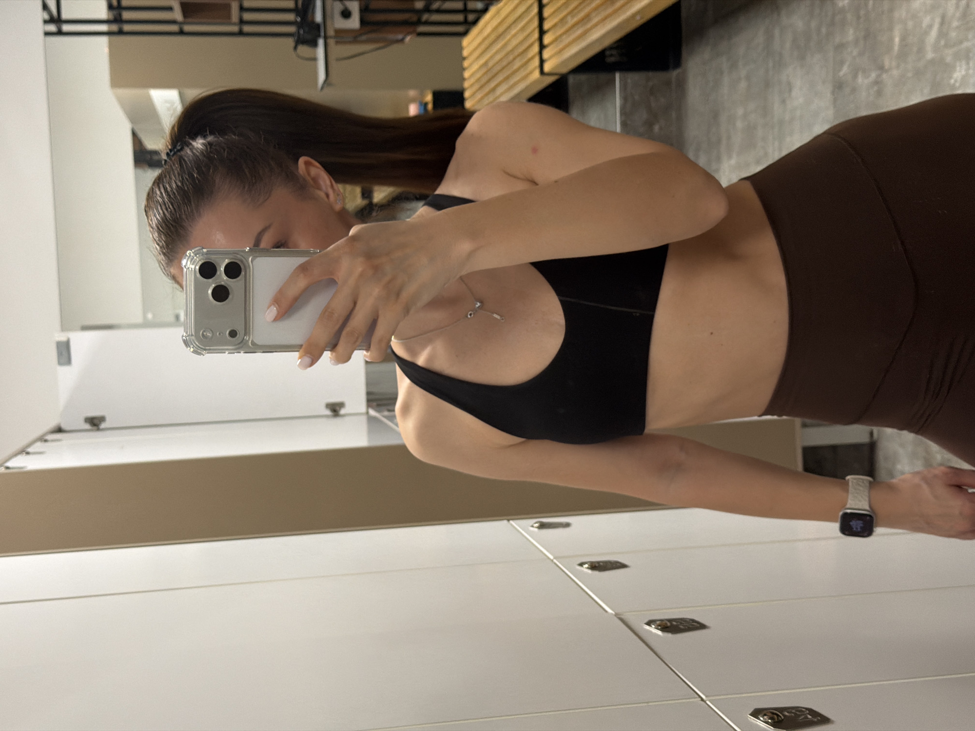 A woman in a black sports bra and brown leggings takes a selfie in a locker room. The locker room is lined with white lockers. She holds a phone with a clear case in her left hand, partially obscuring her face. She wears a silver watch on her left wrist. A wooden bench is visible in the background. It is difficult to ascertain any specific location within Varna, Bulgaria.