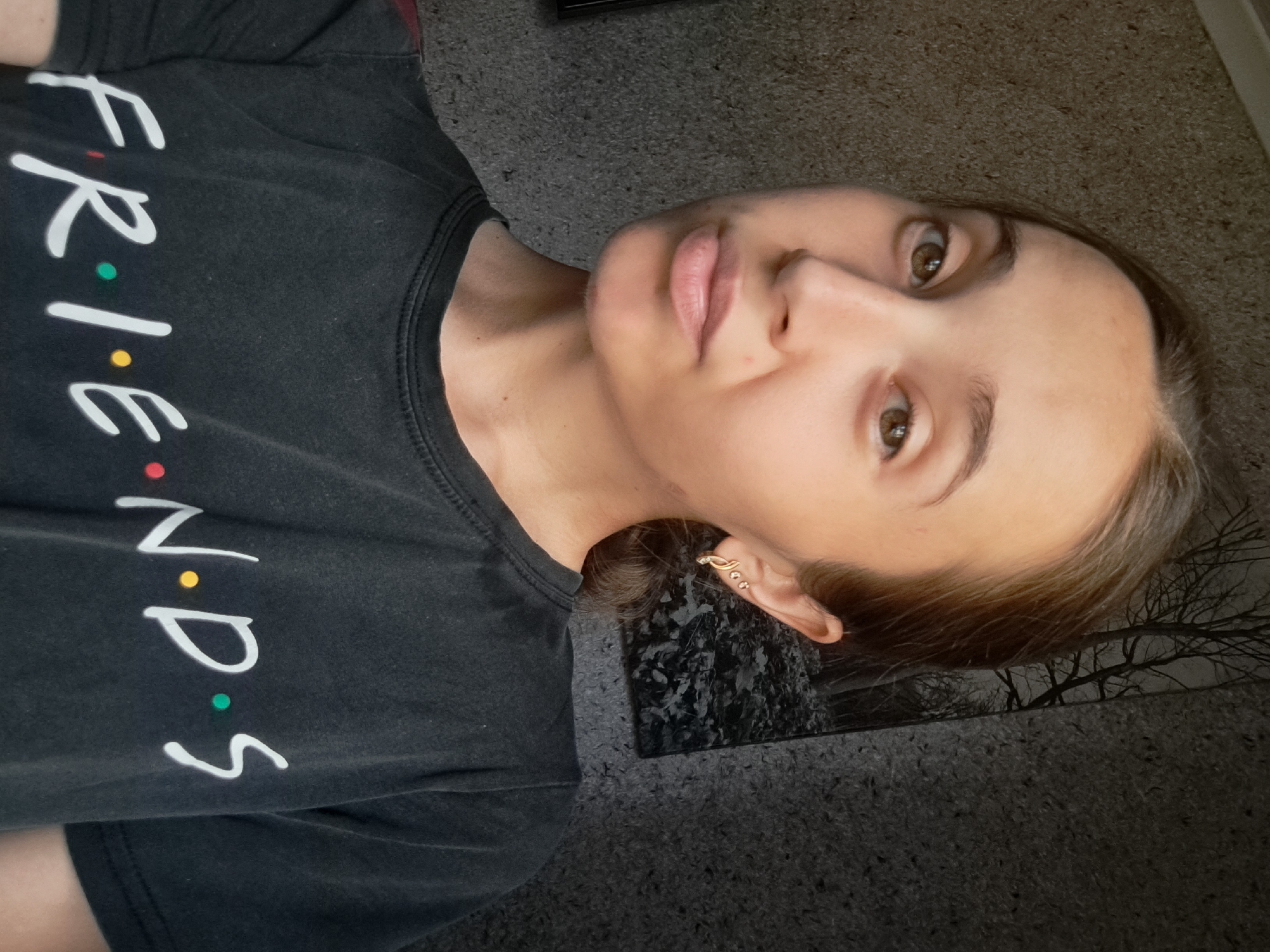 A young woman with brown hair pulled back in a ponytail is looking at the camera. She is wearing a black t-shirt with the word "FRIENDS" printed in white letters, with colored dots representing the "i"s. She has on gold earrings. The background appears to be a textured wall, with a dark, silhouetted image of trees. The lighting suggests the photo was taken indoors during the day. The specific location in Vypasne, Ukraine, is not discernible from the image.