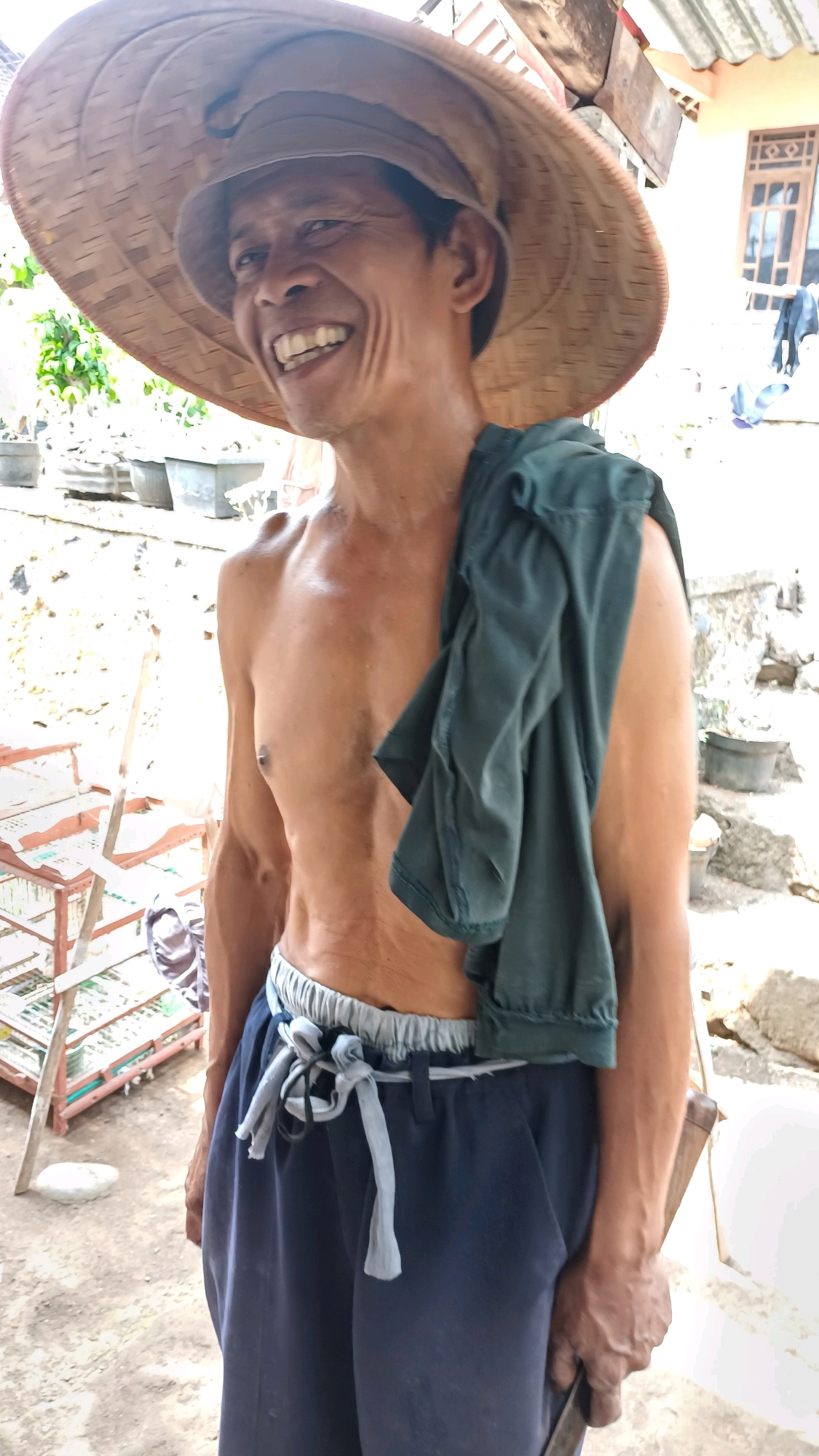 A man in a large, conical straw hat stands outdoors in what appears to be a rural or semi-rural area, possibly in Wonokerso, Indonesia. He is shirtless, revealing his chest and arms, with a dark green t-shirt draped over his right shoulder. He wears dark blue pants with a light gray elastic waistband and a tie. His face is turned to his left, and he is smiling broadly, with his mouth open and teeth visible. His skin is tanned and weathered. In the background, there is a light-colored, rough-textured wall or structure, and what looks like a metal rack or shelving unit with some items on it. Further back, a window with wooden frames is visible on a building. The ground is dirt or gravel.