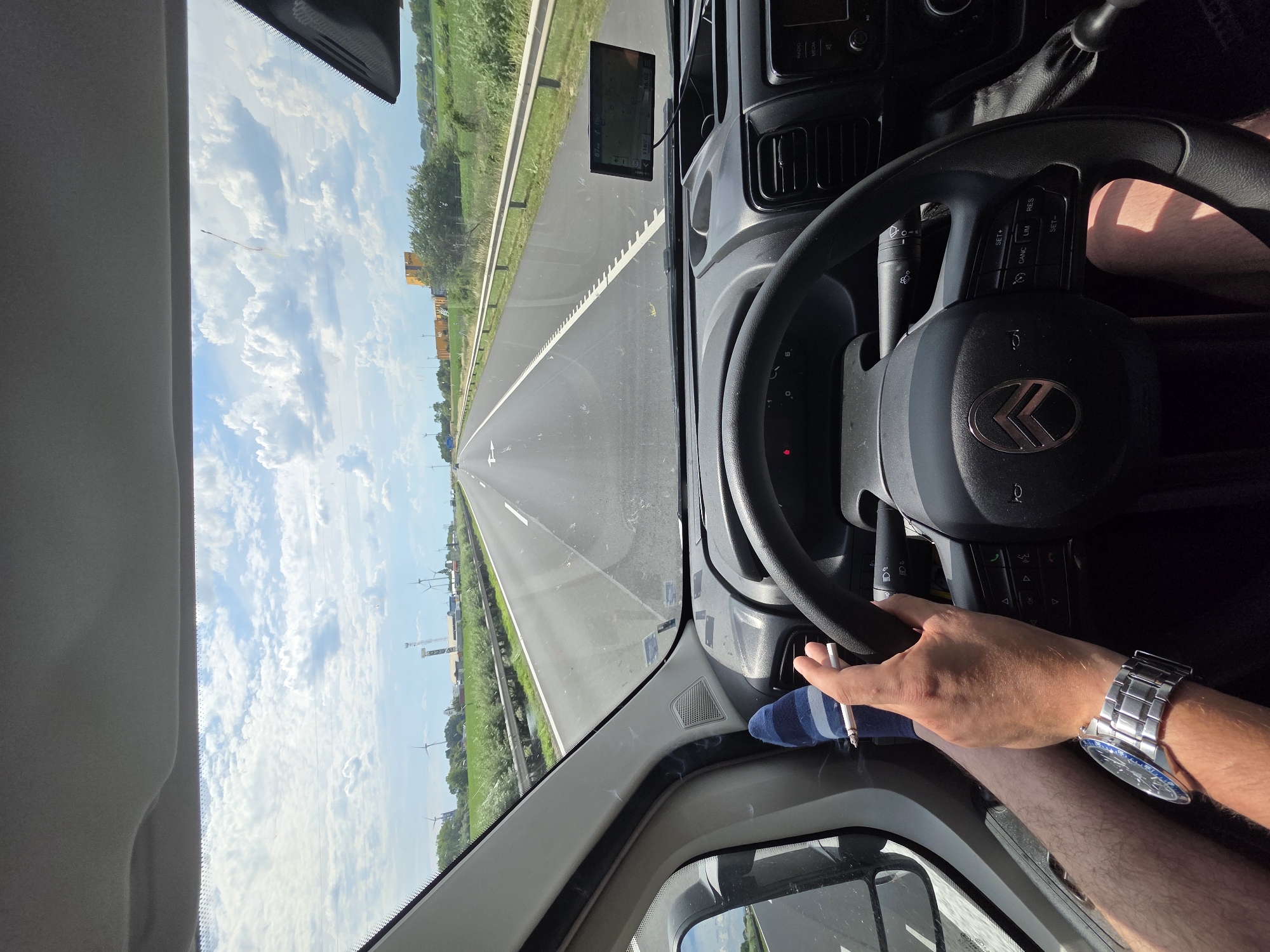 The image depicts the interior of a vehicle on a highway, likely near Kisigmánd, Hungary, given the presence of wind turbines in the distance, a common sight in the region. A person's hands and feet are visible. One hand holds a lit cigarette, and the other rests on the steering wheel, which bears the Citroën logo. A wristwatch is worn on the wrist of the hand holding the cigarette. The feet are clad in striped socks, with one foot resting on the dashboard. The dashboard features a GPS device. The road stretches ahead, with fields and wind turbines visible on either side under a cloudy sky.