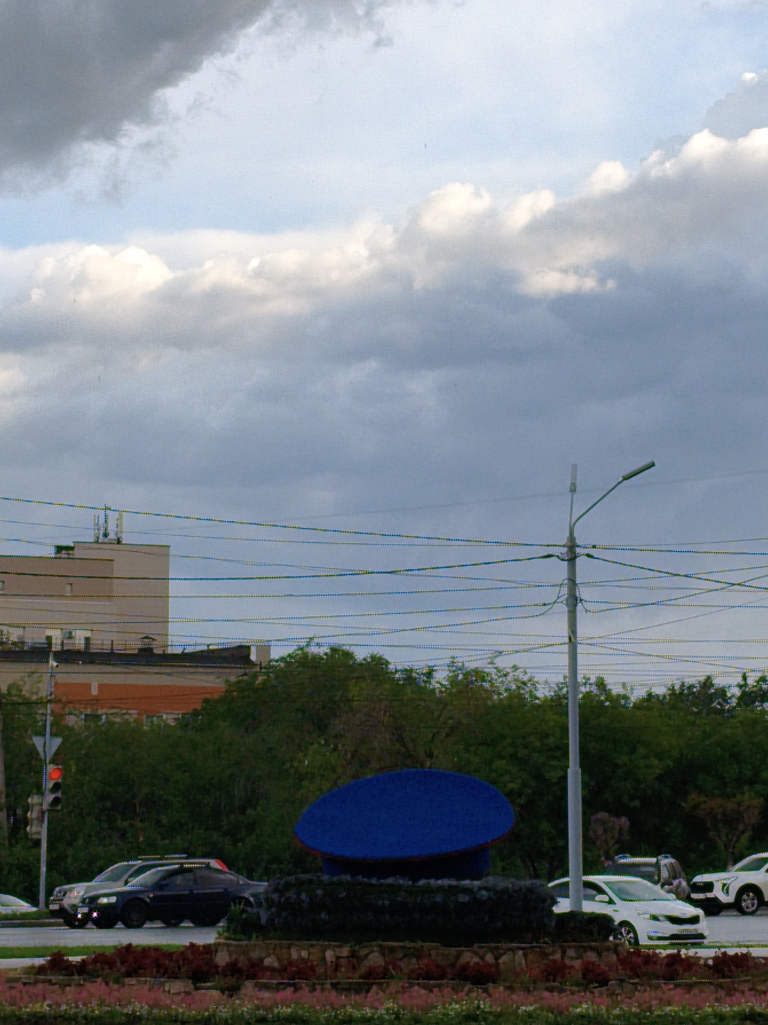 This image depicts an urban scene with a focus on cars and infrastructure against a backdrop of a cloudy sky. In the foreground, a large, bright blue, disc-shaped object sits atop a dark, textured surface, possibly a decorative element or monument. Below this, a flowerbed with pink and red flowers adds a touch of color to the scene.
A street runs through the middle ground, where several cars are visible. A dark sedan and a lighter colored sedan are in motion or stopped at a traffic light, which shows a red signal. Further down the street, a white SUV is also present. The cars suggest a typical day in a city, possibly during rush hour or a period of moderate traffic.
In the background, a multi-story building with a beige facade and darker roofline is seen. Above it, another building with a similar color scheme and what appear to be communication antennas is visible. A prominent streetlight with a single lamp stands tall, accompanied by a complex network of overhead wires that stretch across the frame, indicating the presence of electrical or communication infrastructure. The trees behind the street and in front of the buildings are lush and green, suggesting a time of year between spring and autumn.
The sky is filled with a mix of dark, heavy clouds and patches of brighter, lighter clouds, hinting at overcast or potentially changing weather conditions. The overall lighting suggests it is daytime, possibly late afternoon or early evening, given the diffused light. There are no people clearly visible in the frame. The scene is likely set in an urban environment, as indicated by the buildings, street, traffic, and power lines. The presence of a traffic light and multiple vehicles points to a functional road network.