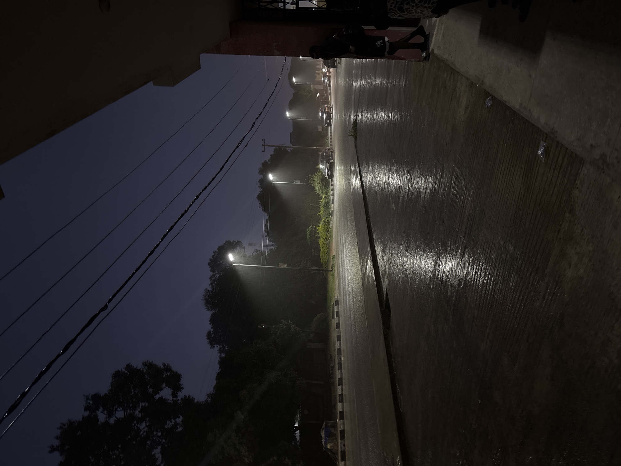 The image depicts a street scene at dusk in Jos, Nigeria, during a rainfall. The wet asphalt road reflects the glow of streetlights, with several cars driving by. Trees line the left side of the road, and a building with a red facade is visible on the right. Two individuals are standing by the building on the right side of the frame, partially obscured. The sky is dark and overcast, suggesting the continuation of the rain. Small plastic-wrapped items are visible on the pavement near the building. Overhead power lines crisscross the sky.