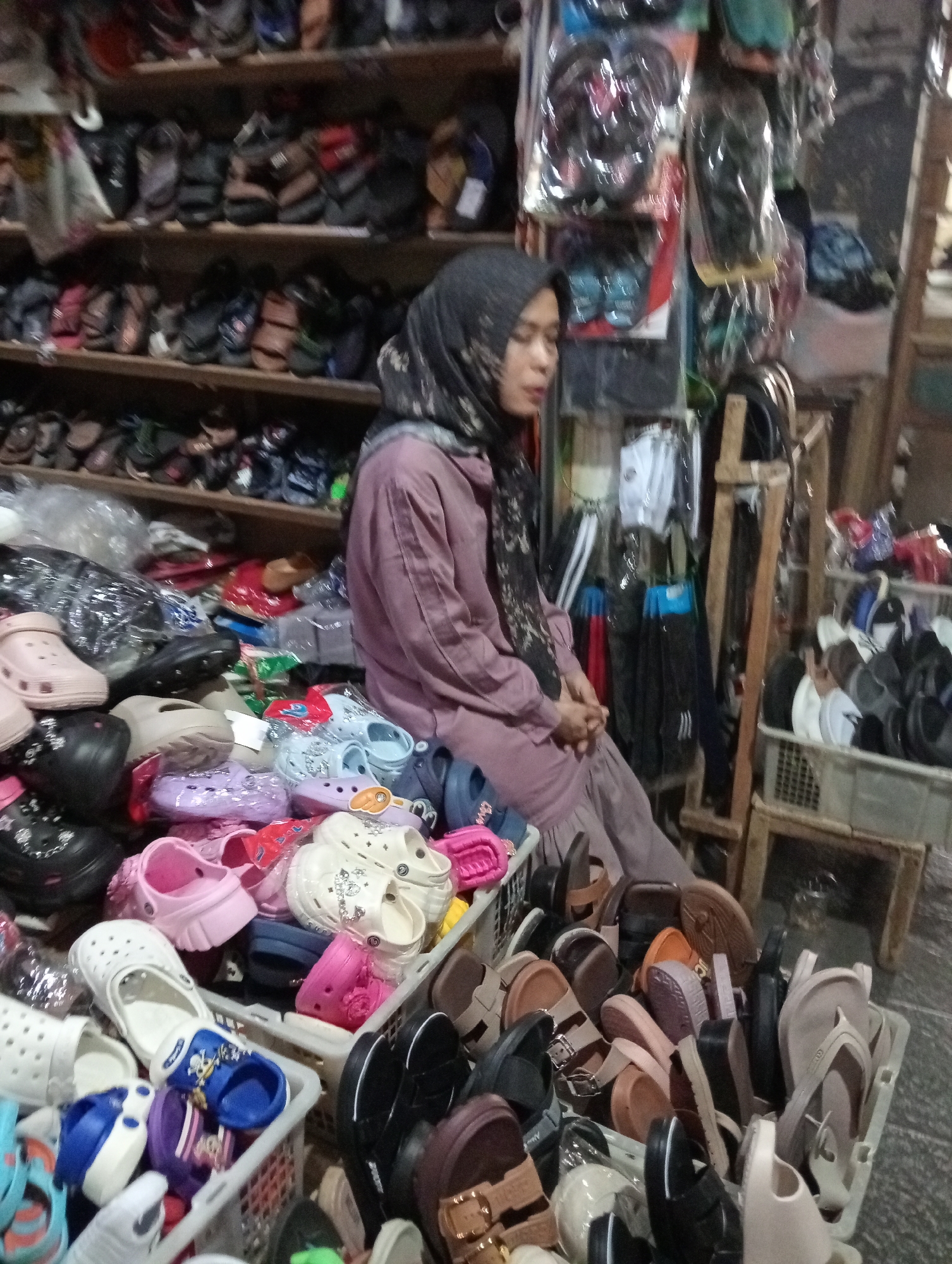 A woman wearing a hijab and a mauve dress sits in front of a market stall filled with shoes. The stall has shelves stocked with various types of footwear, including sandals, clogs, and flip-flops. In the foreground, several baskets overflow with more shoes, primarily clogs in a variety of colors like pink, white, and light blue. The woman is looking to her left, with her hands resting in her lap. The overall environment appears to be an indoor market. Based on the context provided, this scene is located in the city of Grabag, Indonesia.