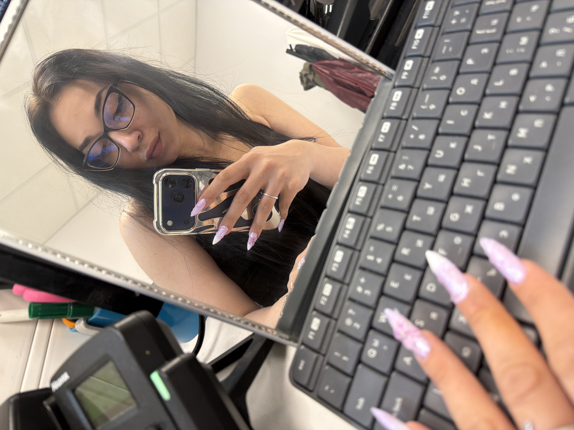 In Stara Zagora, Bulgaria, a woman is taking a selfie in a mirror. She is wearing glasses and has long dark hair. She is holding a phone in her right hand, which is resting on a keyboard. Her fingernails are long and painted with a light purple glitter. In the reflection of the mirror, a device with a screen is visible to the left, and some colorful bottles and a green marker are in a blue holder behind it. To the right of the mirror, some clothing items hang on a rack. The scene appears to be indoors, possibly in an office or salon.