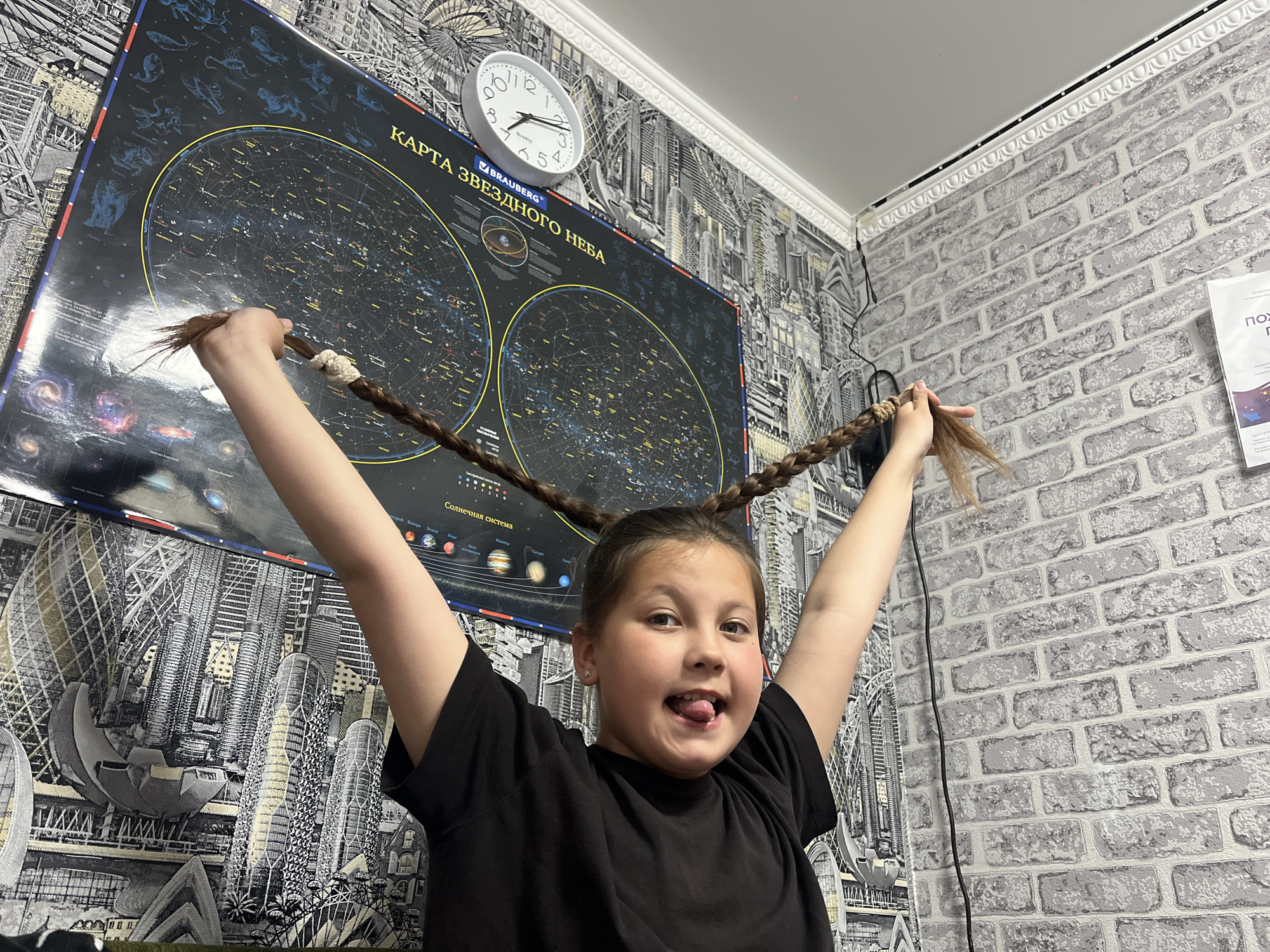 A young girl is standing with her arms raised, holding the ends of her braids. She is wearing a black shirt and sticking her tongue out. Behind her is a wall with a black and white cityscape wallpaper and a poster of the "KAPTA ЗВЕЗДНОГО НЕБА" (Starry Sky Map) along with a clock. The other wall has a light gray brick pattern wallpaper. The scene takes place indoors. There is no mention of the city of Novopokrovskaya, Russia, in the image.