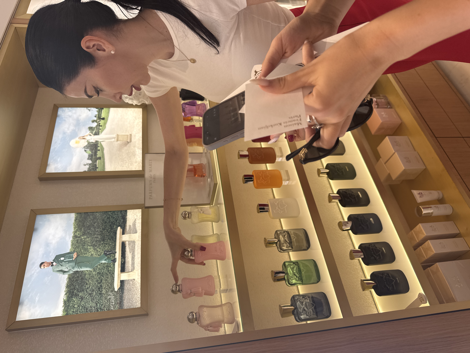 The image depicts an interior scene, likely a retail environment, featuring a display of perfumes. A woman with dark hair pulled back is in the foreground, wearing a white t-shirt and a red skirt. She is pointing at a pink perfume bottle on a shelf, engaging in what appears to be a product selection or discussion. Her right hand holds a small white card with text visible, and her left hand is near a smartphone. Another person's hands are visible in the lower right, holding sunglasses and a portion of the white card. The background displays several shelves stocked with various perfume bottles in different colors and shapes, along with framed artwork depicting fashion-inspired imagery. The lighting is warm, illuminating the products and creating a polished aesthetic. The location is specified as Germasogeia Municipality, Cyprus.