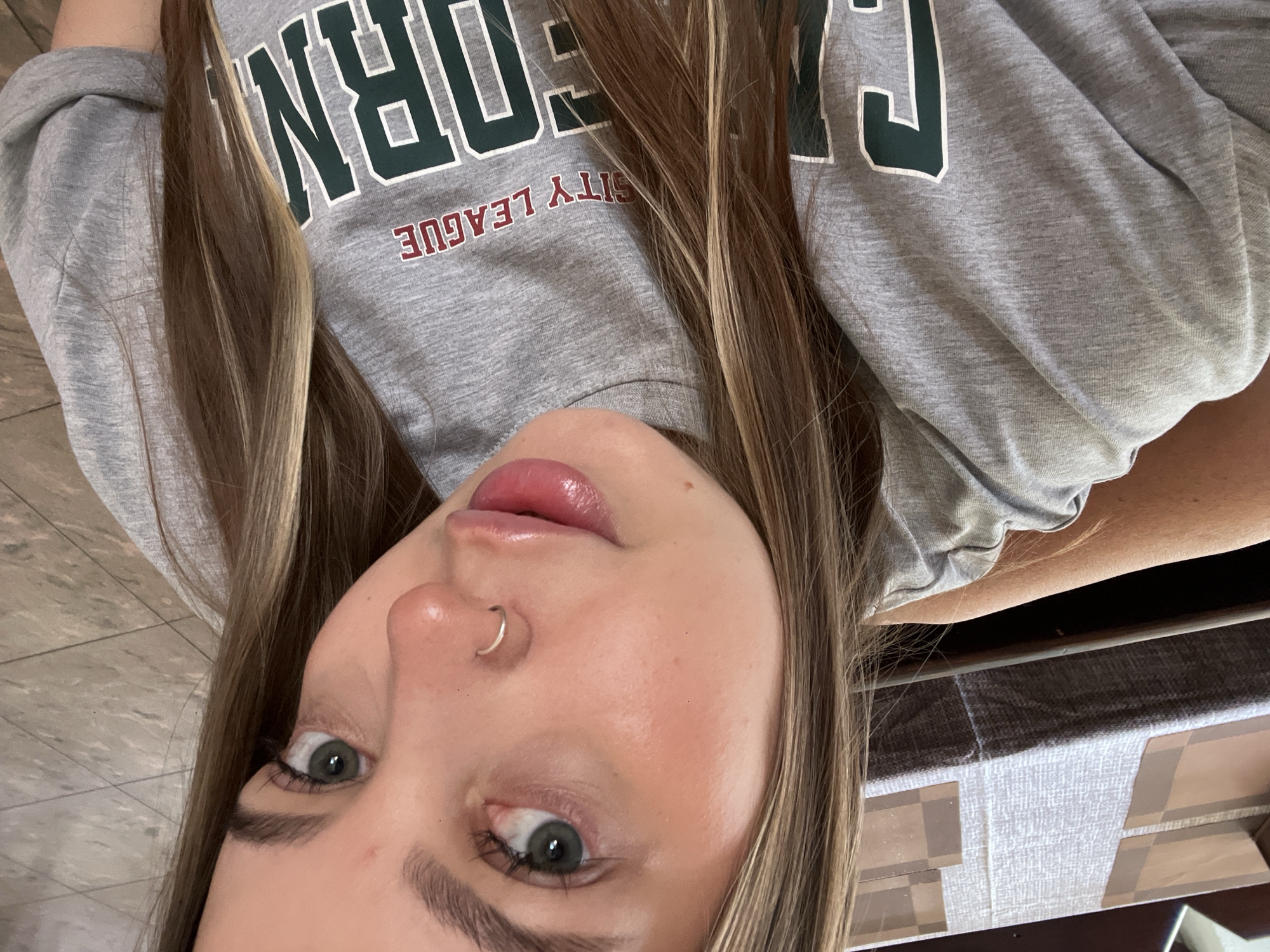 The image features a close-up, high-angle selfie of a young female individual in an indoor setting within Novopokrovskaya, Russia. She has long, straight brown hair with blonde highlights, parted centrally, cascading over her shoulders. Her gaze is directed towards the camera, revealing green eyes with distinct eyelashes and well-defined, dark eyebrows. A small, circular, silver-colored hoop piercing is visible in her left nostril. Her lips are full, glossy, and tinted a rosy pink, slightly parted. She wears a light grey crew-neck top, likely a t-shirt or sweatshirt, with the sleeves rolled up. The garment displays partial text: "SITY LEAGUE" in maroon letters, and the initial letters "CALI" of a larger word, possibly "CALIFORNIA," rendered in dark green with a white outline. The background on the upper left shows a table covered by a light-colored, textured fabric runner, overlaid with two rectangular, light brown placemats. The lower right background reveals a light grey tiled floor with visible grout lines.